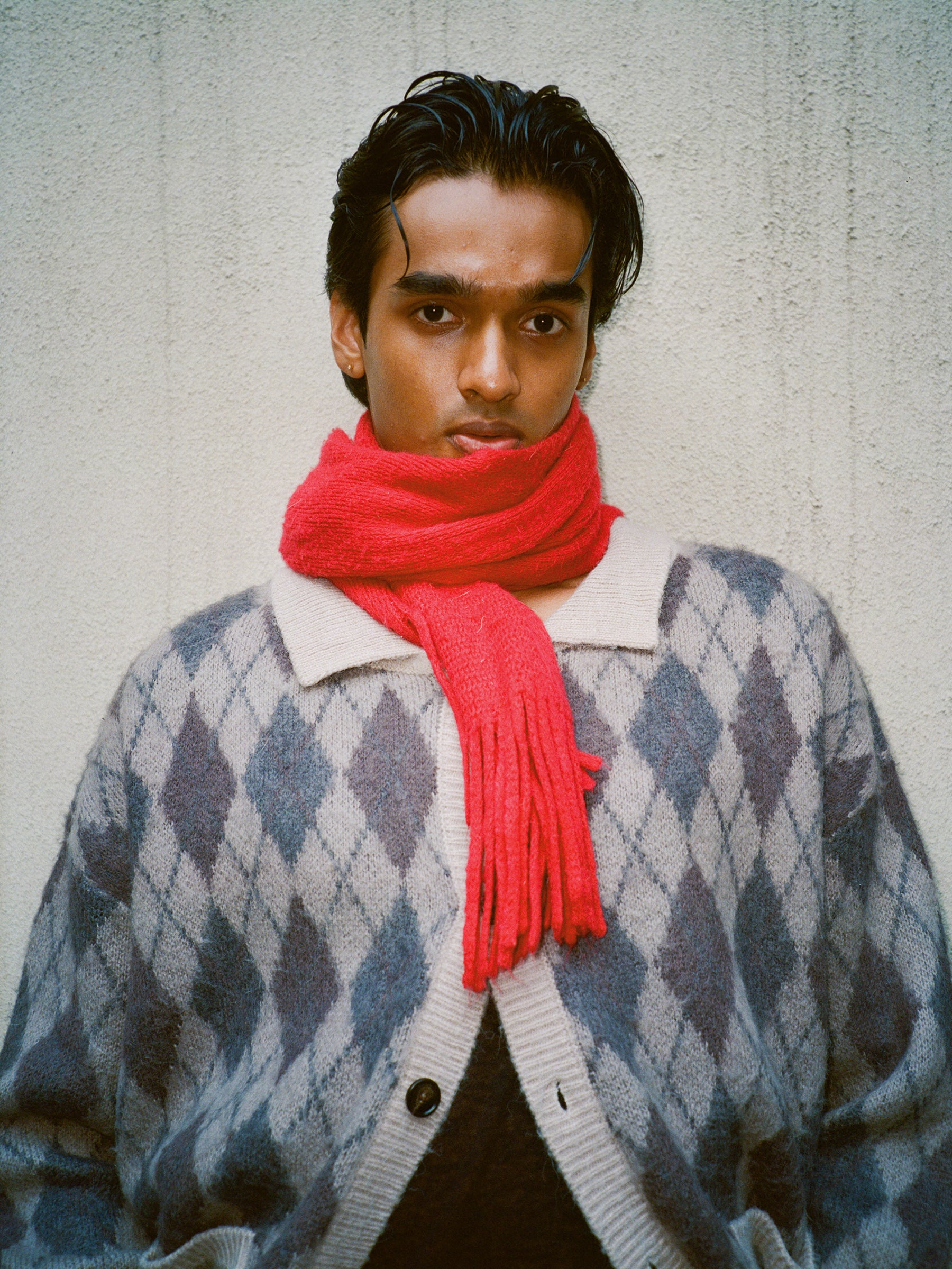A person wearing the FOUND Argyle Cardigan in grey and white paired with a bright red scarf stands against a textured beige wall, looking directly at the camera.