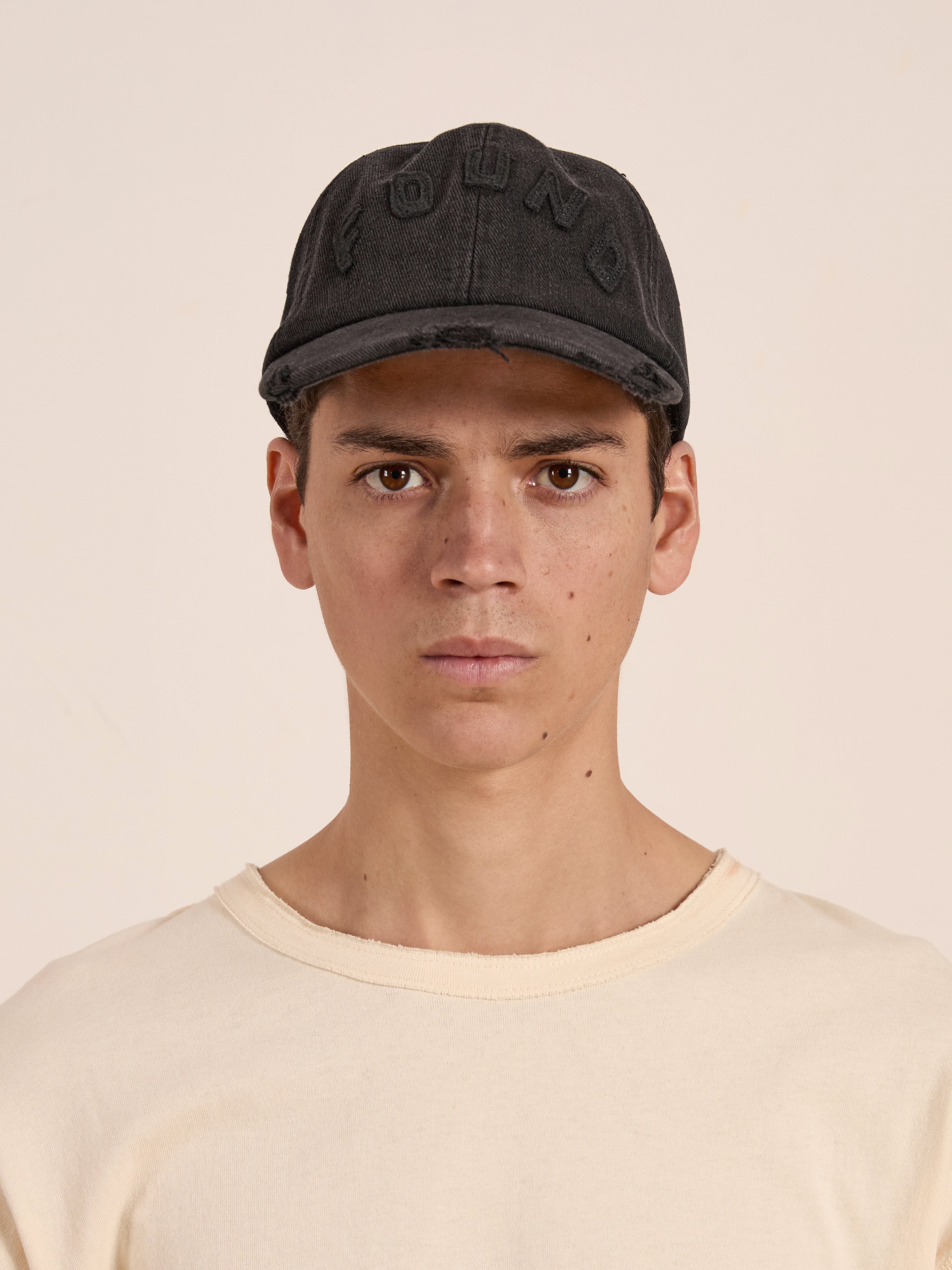 A person in a beige shirt and a Black Felt Patch Cap by FOUND, with "FOUND" stitched on it and a distressed brim, looks at the camera against a plain background.