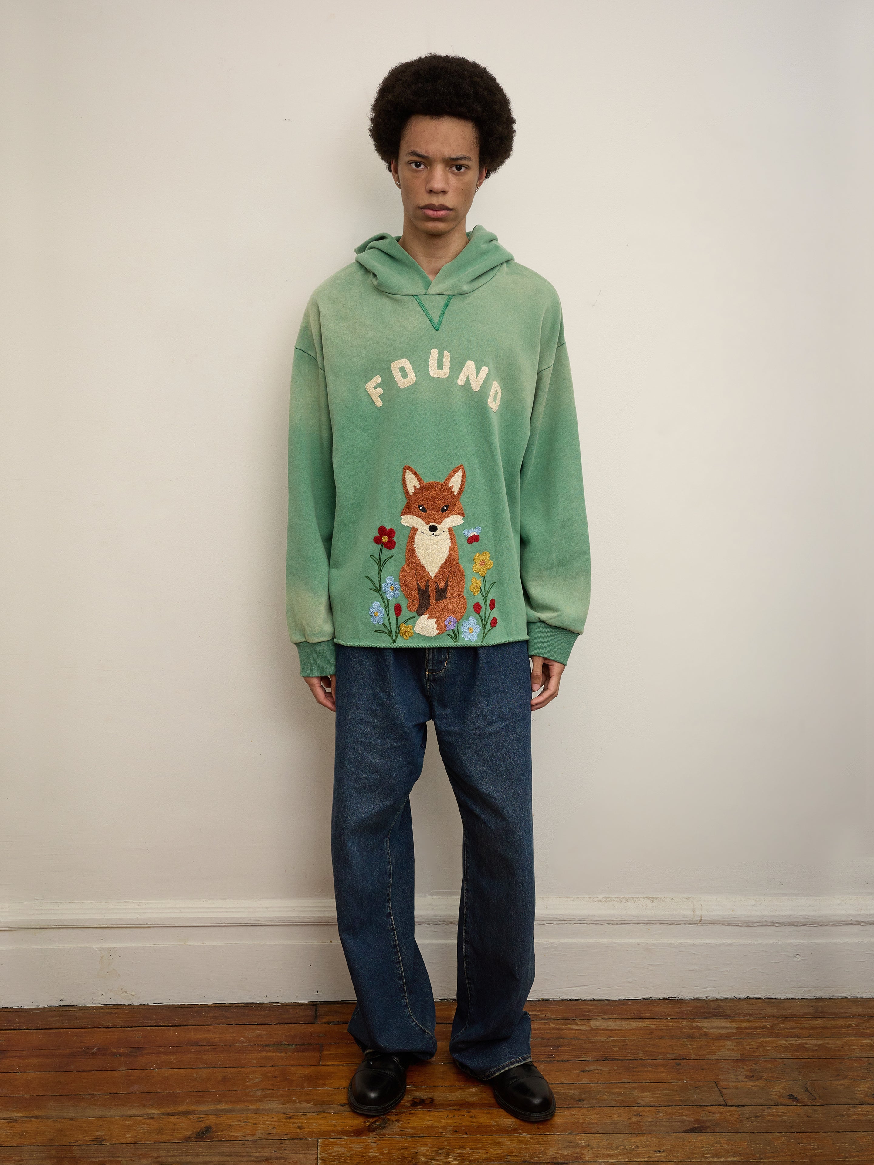 A person stands against a plain wall, wearing a green Embroidered Wildlife Hoodie by FOUND with a fox graphic, styled with blue jeans and black shoes on a wooden floor.