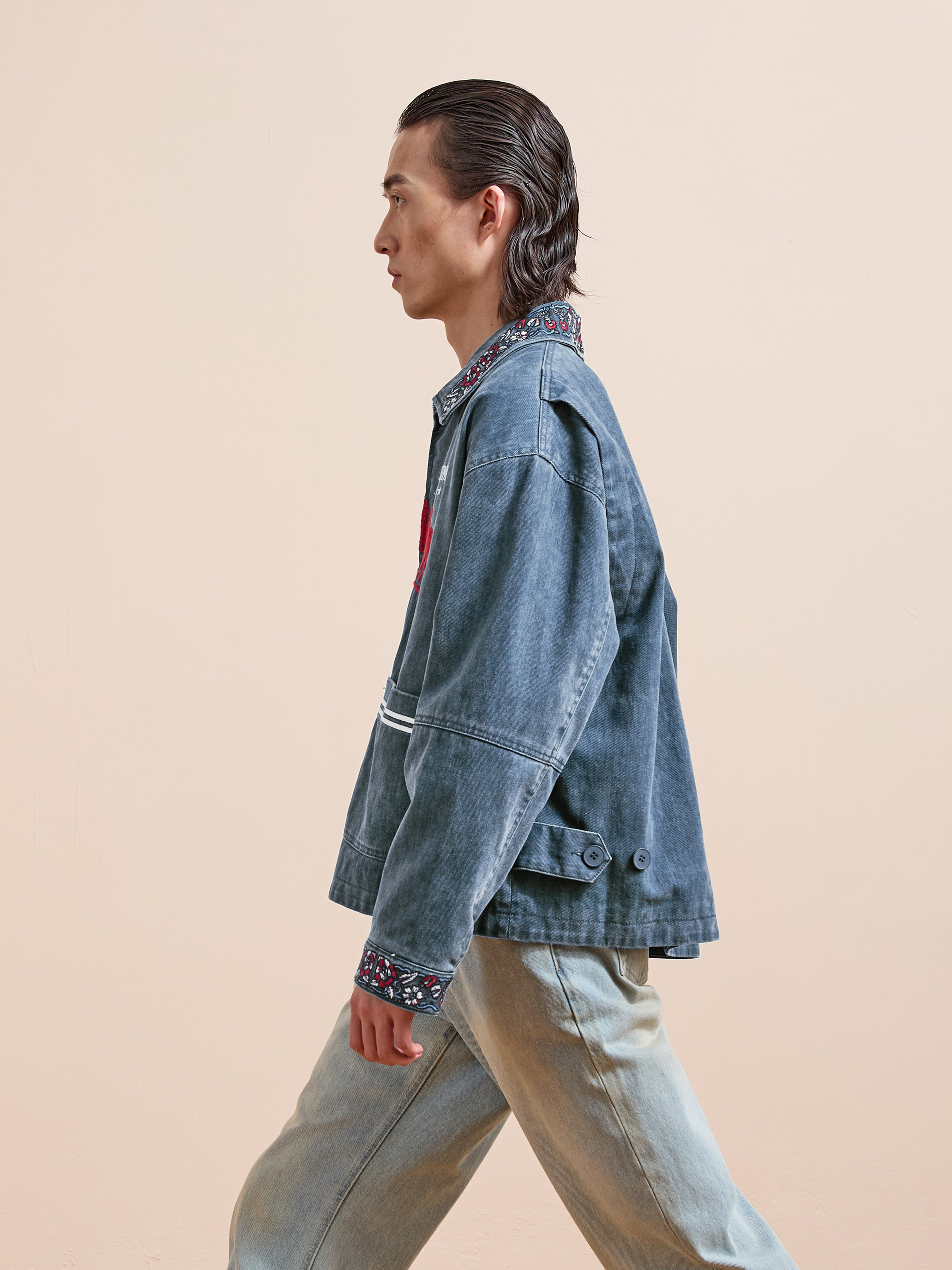 A person with slicked-back hair, in a Found Jockey Club Embroidered Jacket and light unisex relaxed-fit jeans, walks in profile against a plain beige background.