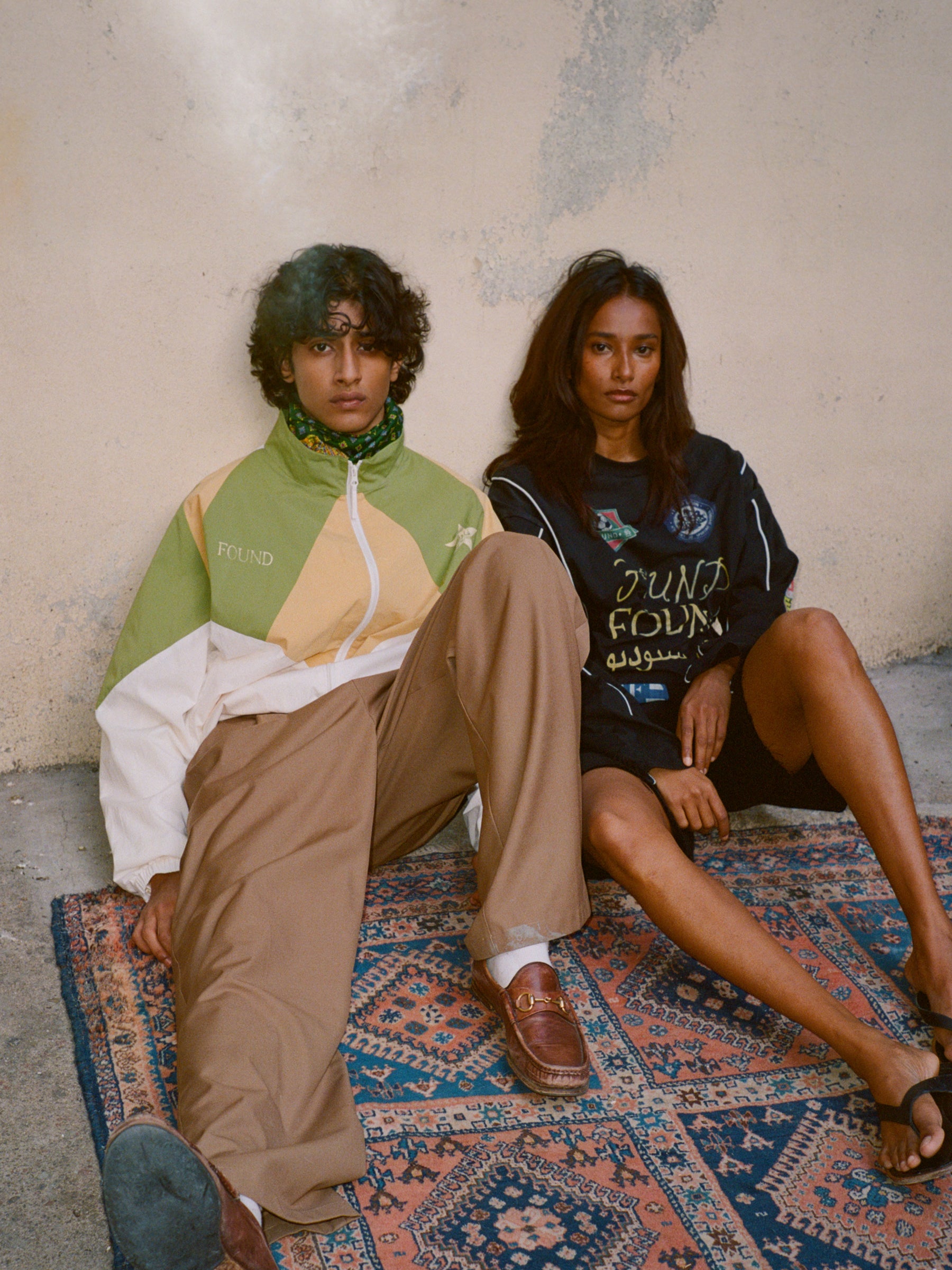Two people sit against a wall on a patterned rug. The person on the left wears a FOUND Legacy Cricket Track Jacket, while the person on the right wears a dark long-sleeve shirt.