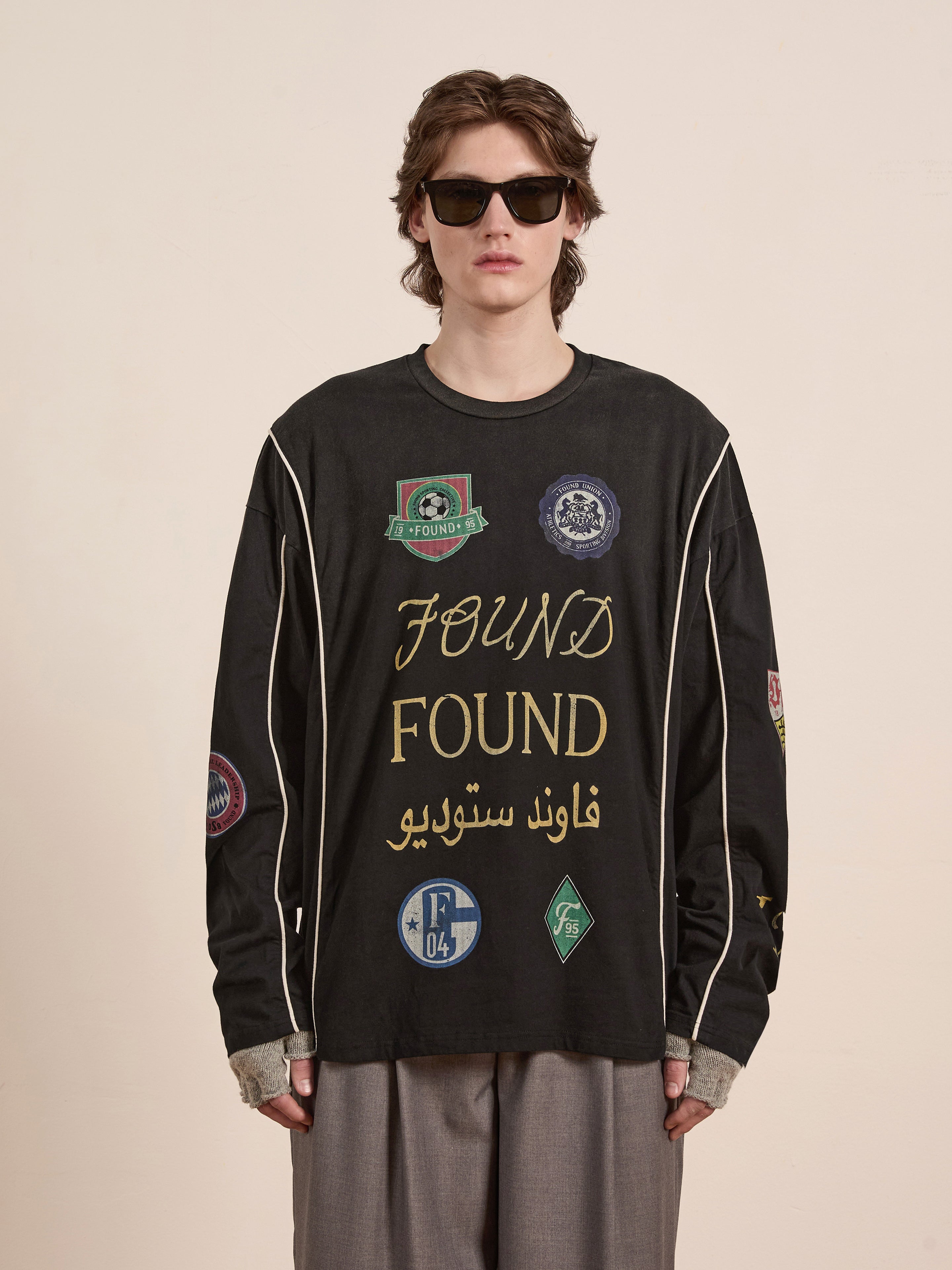 Wearing the unisex Multi Language LS Jersey by FOUND, a person is styled in casual sunglasses and a black long-sleeve shirt featuring various patches and "FOUND" repeated, creating a retro look against a plain backdrop.