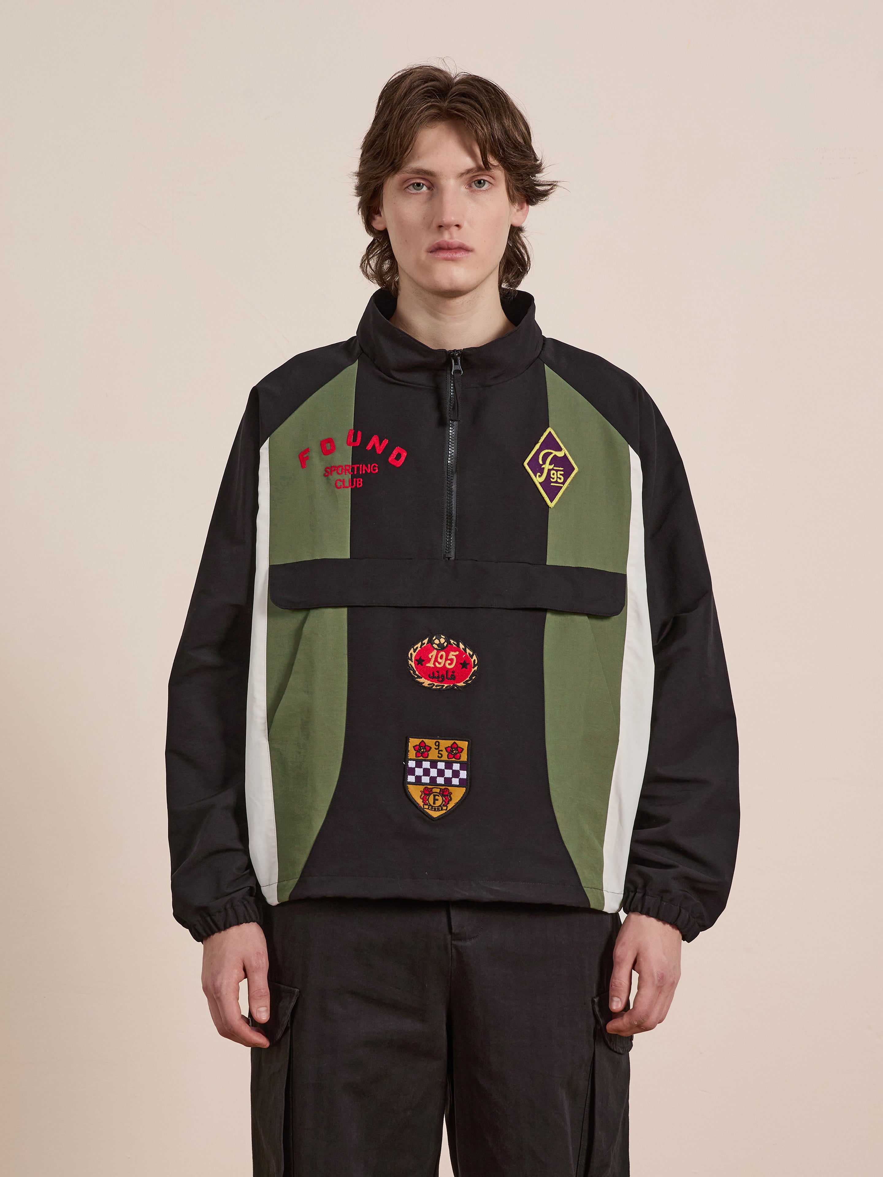 A person wearing FOUND's black and green Sporting Club Pullover Jacket with patch designs stands against a plain background, exuding a vintage aesthetic.