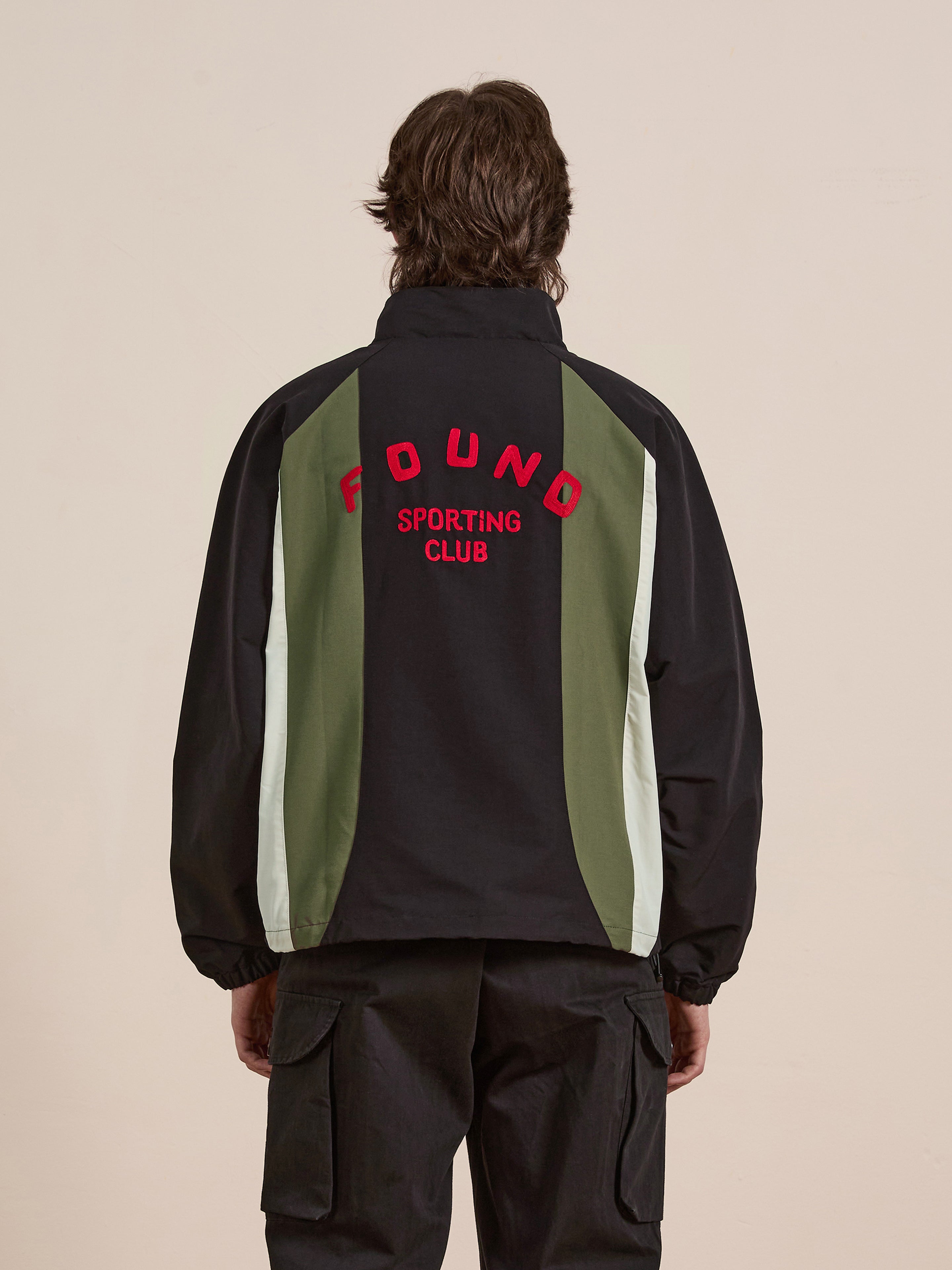 A person is wearing the FOUND Sporting Club Pullover Jacket, which has "FOUND SPORTING CLUB" on the back and black, green, and white panels for a vintage look.
