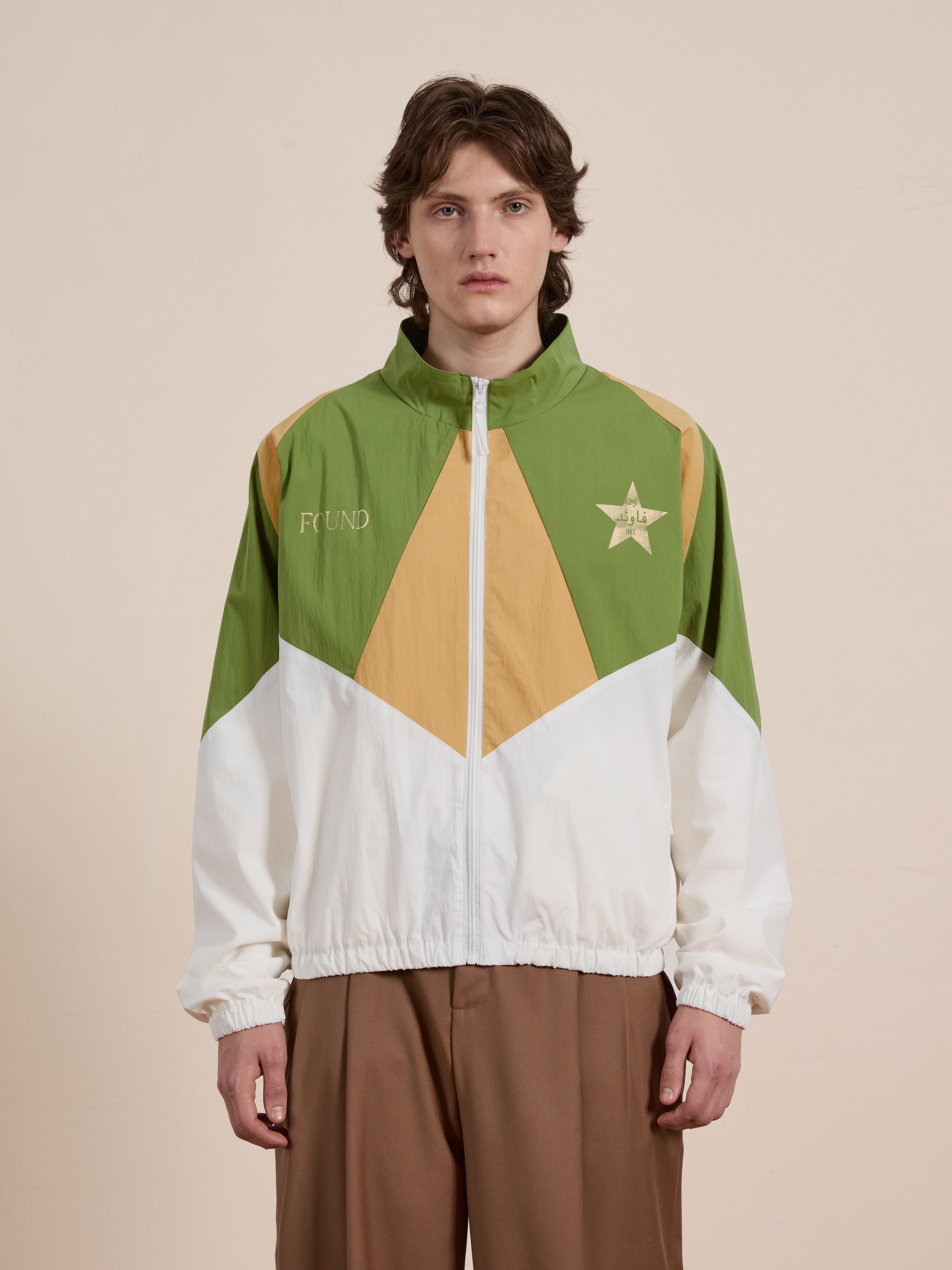 A person models the FOUND Legacy Cricket Track Jacket, featuring green, gold, and white colors with a star emblem. Paired with brown pants against a plain background, it exudes a vintage athletic vibe.