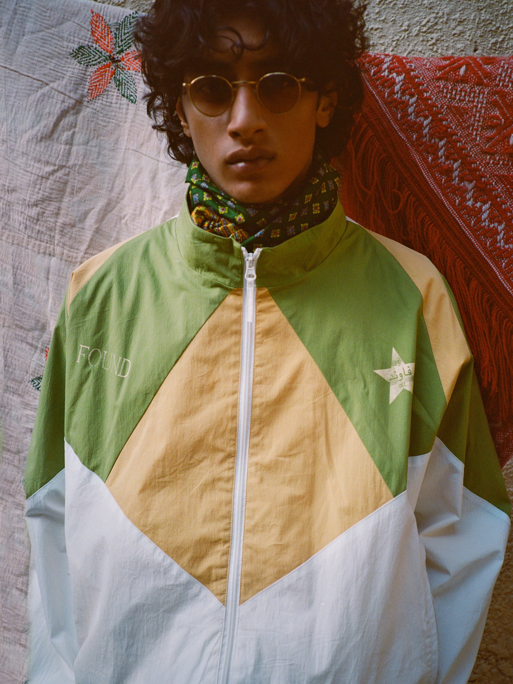 A curly-haired person wearing sunglasses and the FOUND Legacy Cricket Track Jacket stands in front of textured cloths.