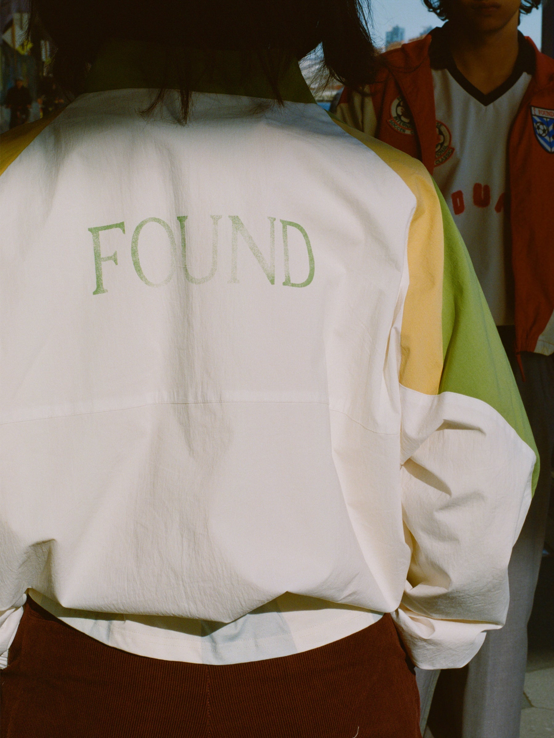 Sporting a white Legacy Cricket Track Jacket by FOUND, a person channels vintage athletic style with "FOUND" on the back, as someone in a sports jersey enhances the retro vibe.