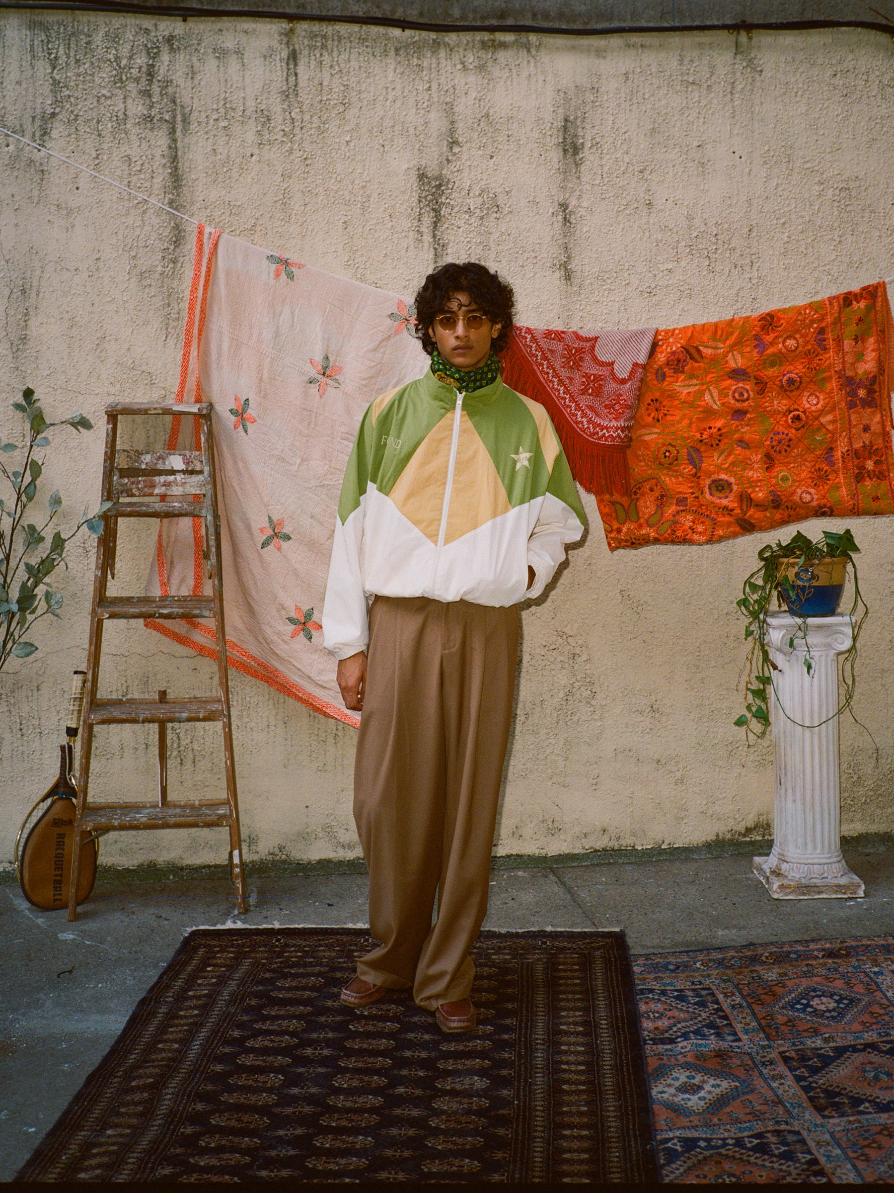 A person in vintage style wears the Legacy Cricket Track Jacket by FOUND, standing before a textured wall embellished with colorful fabrics, beside a rustic ladder and a stringed instrument on the floor.