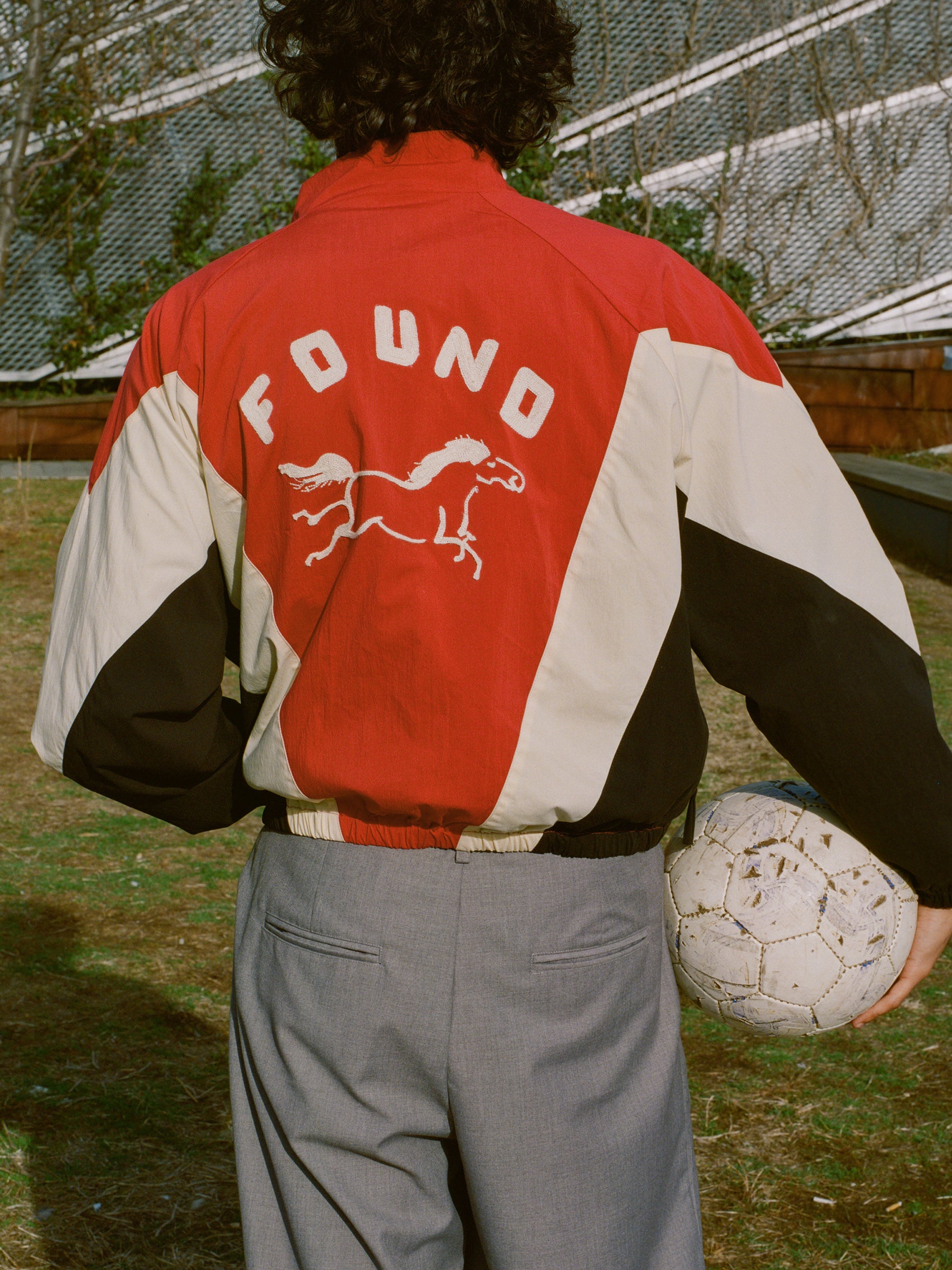 A curly-haired person wears a Pine and Rose Track Jacket by FOUND, featuring "FOUND" and a horse logo. They hold a worn soccer ball while standing on grass with a building in the background.