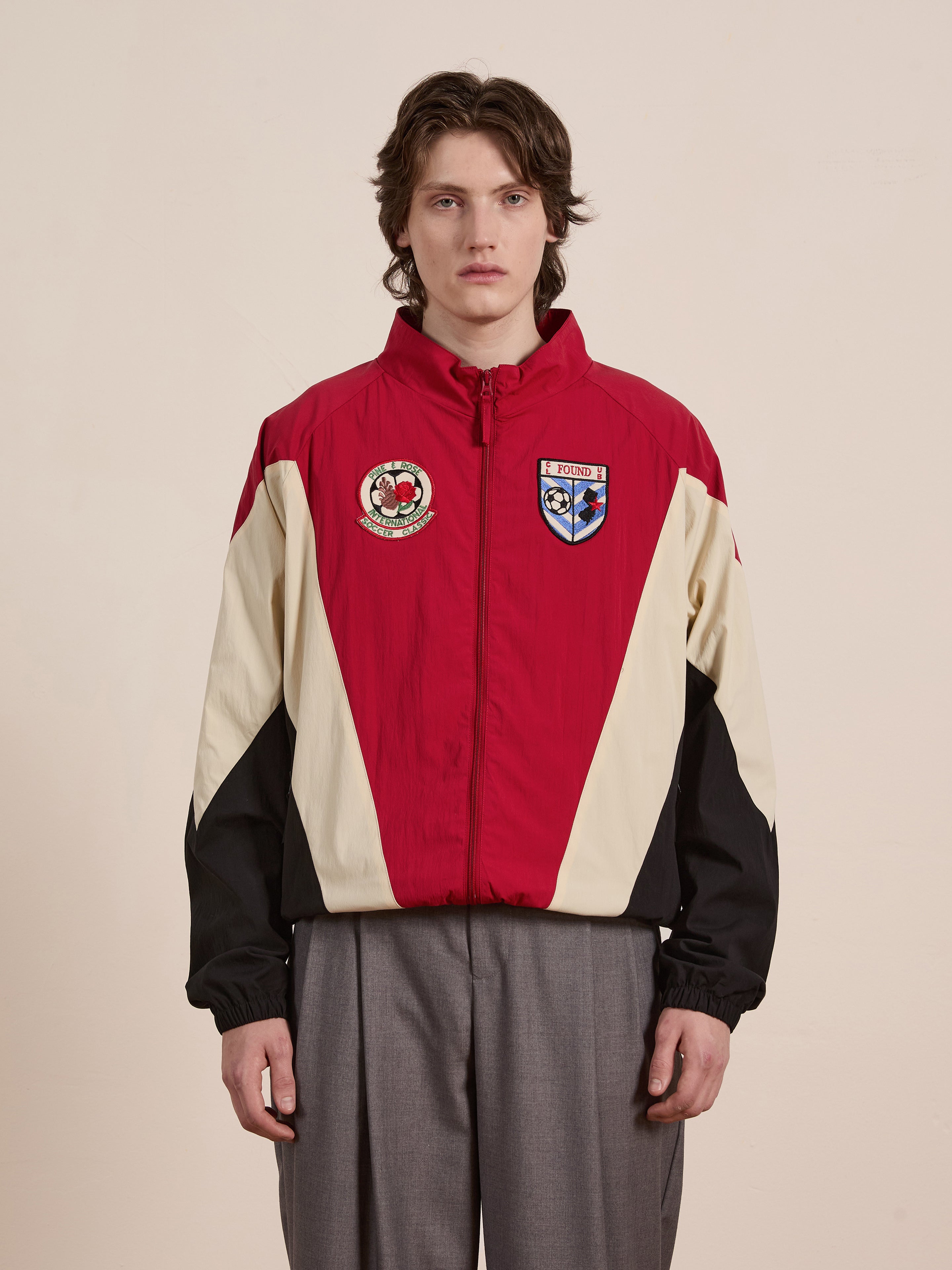 A person poses against a plain background wearing the chic, unisex FOUND Pine and Rose Track Jacket with red, black, and beige patches, paired with gray pants. This piece seamlessly blends form and function.