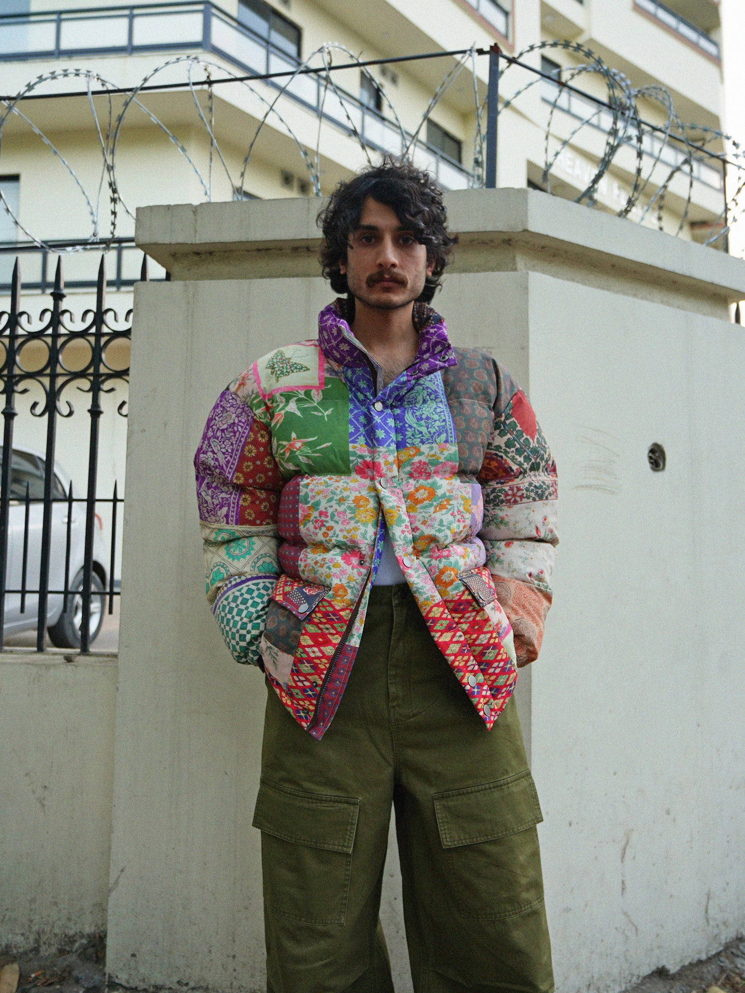 A man wearing a colorful jacket, the Found Gardenia Tapestry Puffer Jacket, standing next to a fence.