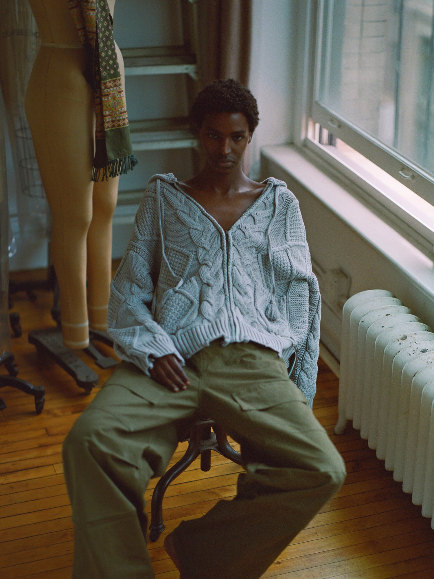 A man wearing a chunky knit Zip Up Distressed Cable Knit Hoodie by Found sitting on a chair in front of a window.