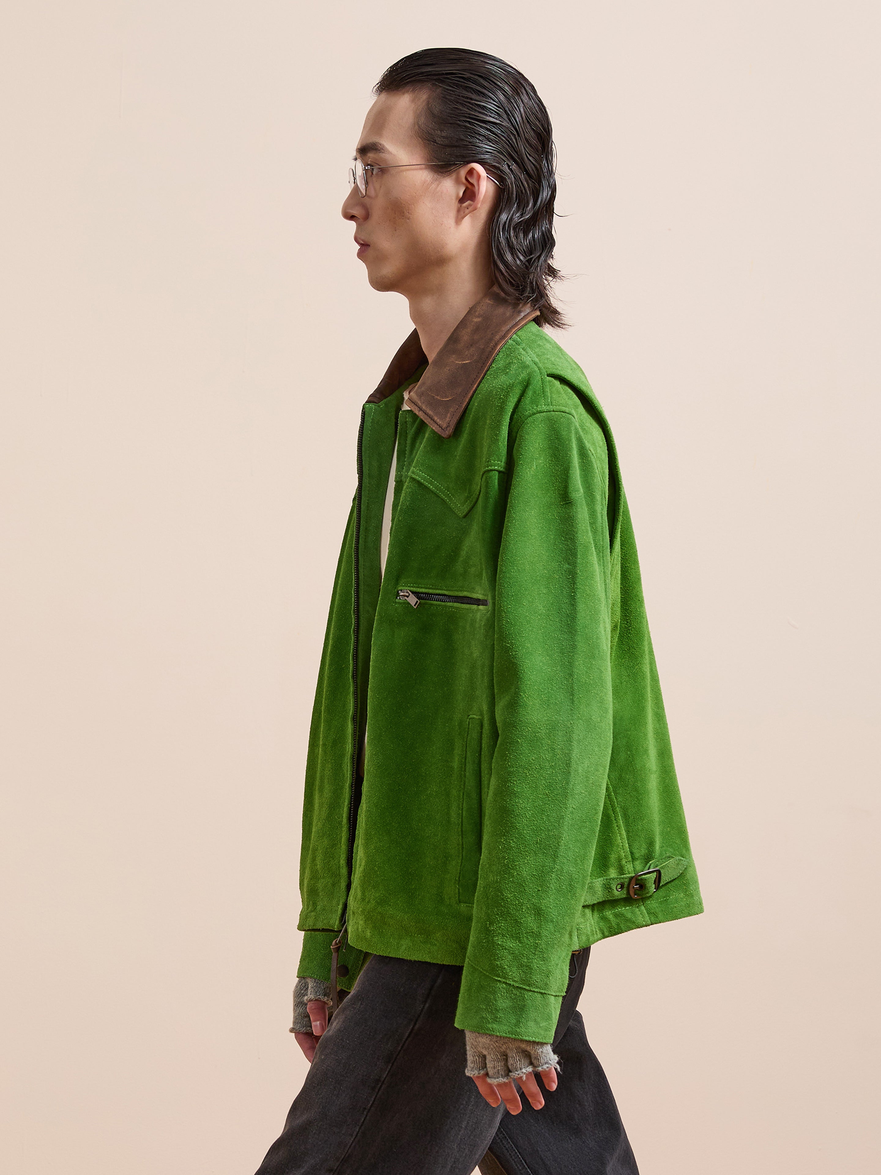 A person in profile wears the FOUND Suede Western Barn Jacket in bright green with a distressed leather collar, paired with gray fingerless gloves, black pants, and glasses, standing against a plain beige background.