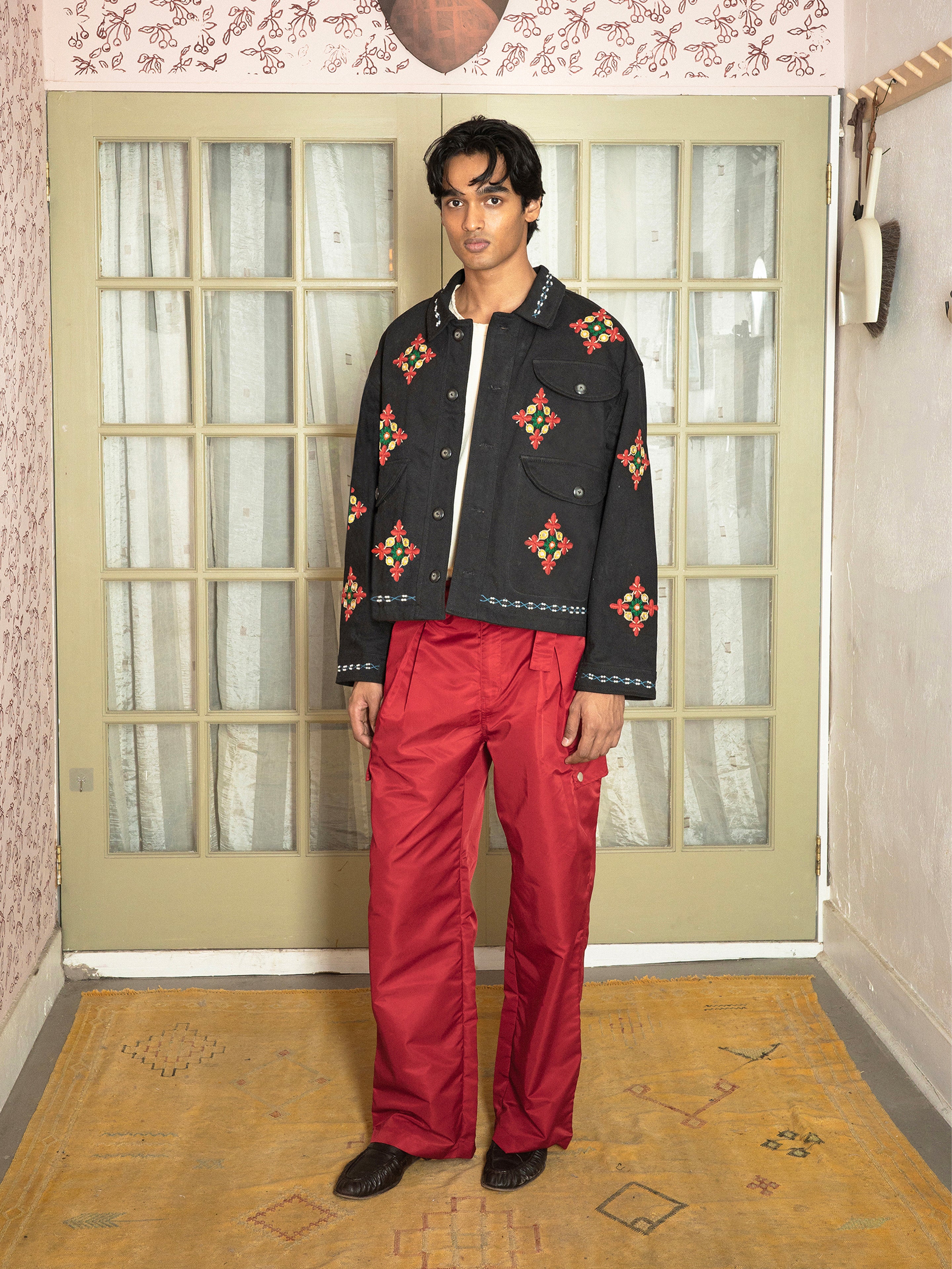A person stands indoors in front of glass-paned double doors, wearing Found’s Mirror Ornament Hand Embroidered Work Jacket with red and green patterns, paired with shiny red pants.