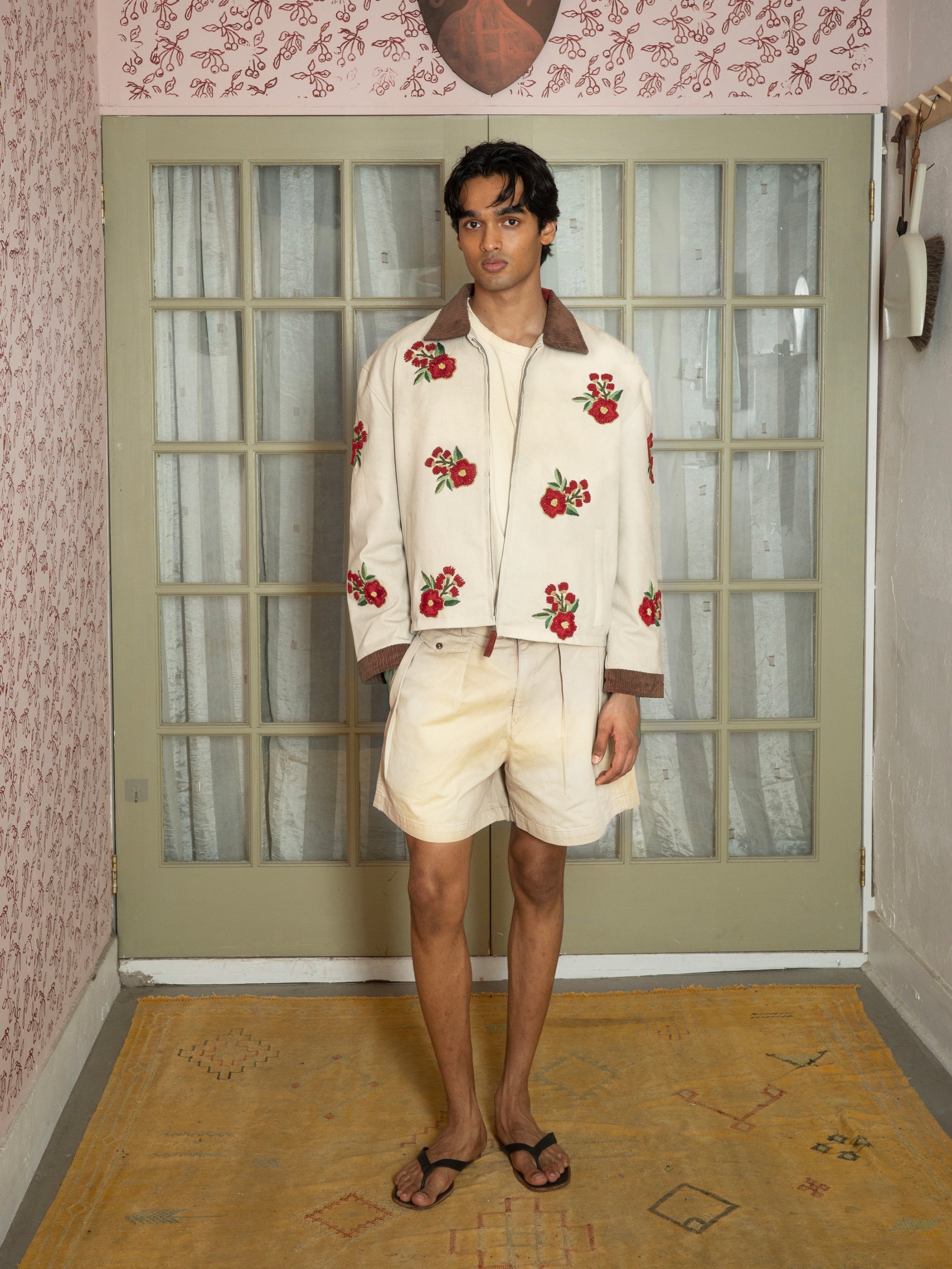 A man indoors wears the Found Floral Hand Embroidered Work Jacket, inspired by vintage South Asian needlework, styled with cream shorts and flip-flops. He stands on a patterned rug in front of glass doors.