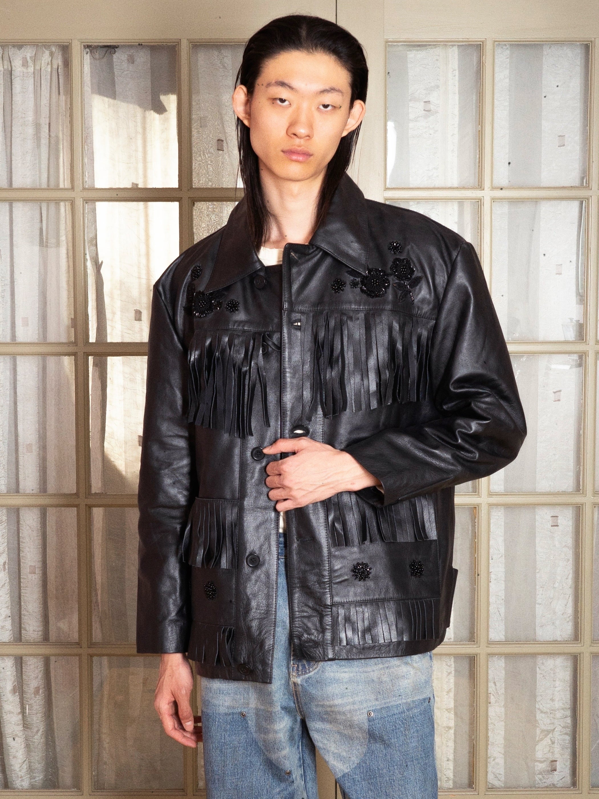 A person models the FOUND Hand Embroidered Leather Fringe Jacket, made of genuine cowhide, in front of glass-paneled doors, holding one side of the unisex jacket and looking at the camera.