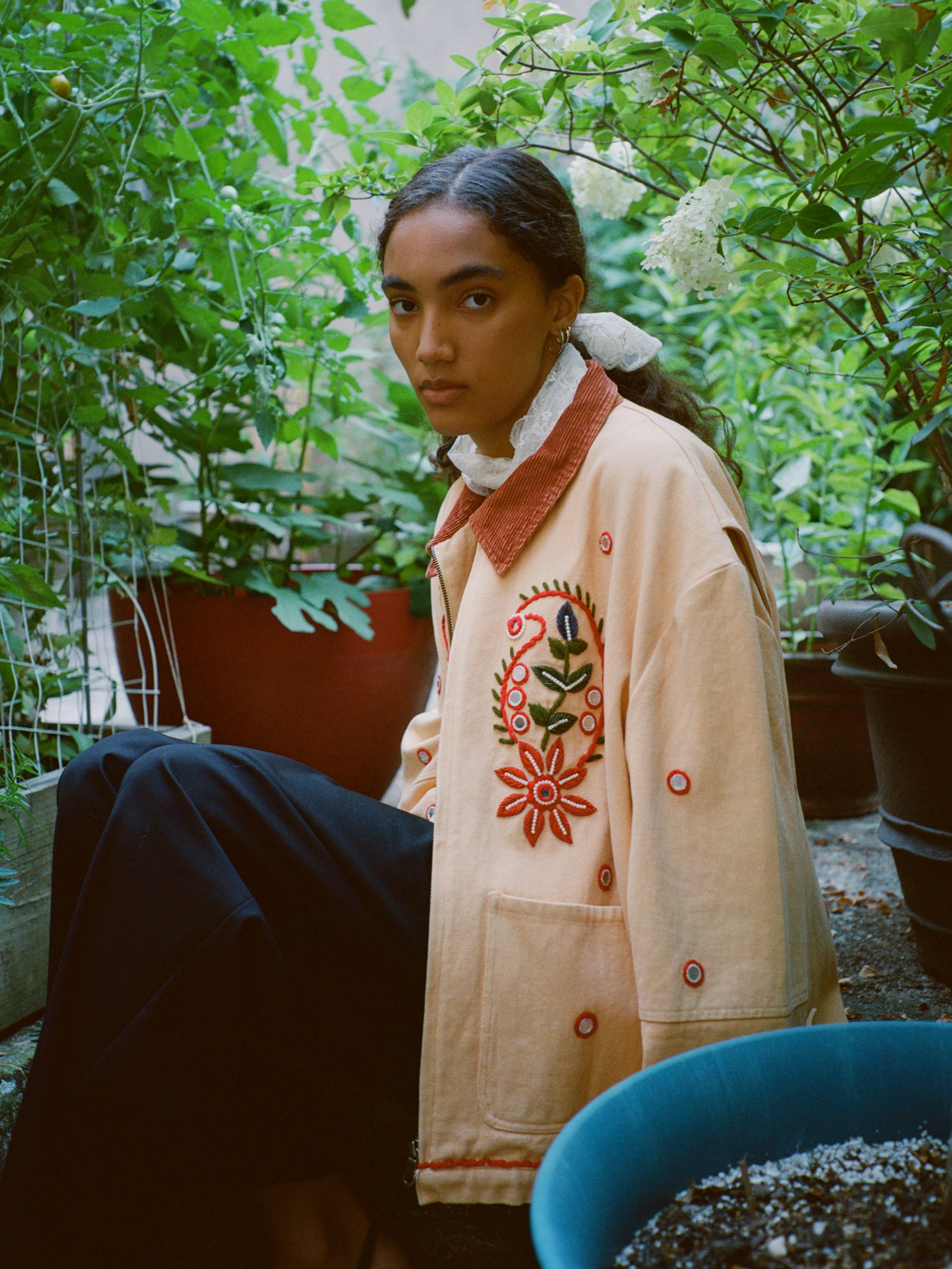 A person with long dark hair sits among green plants, wearing the Found Paisley Motif Hand Embroidered Work Jacket, featuring artisan craftsmanship and colorful floral details, paired with black pants.