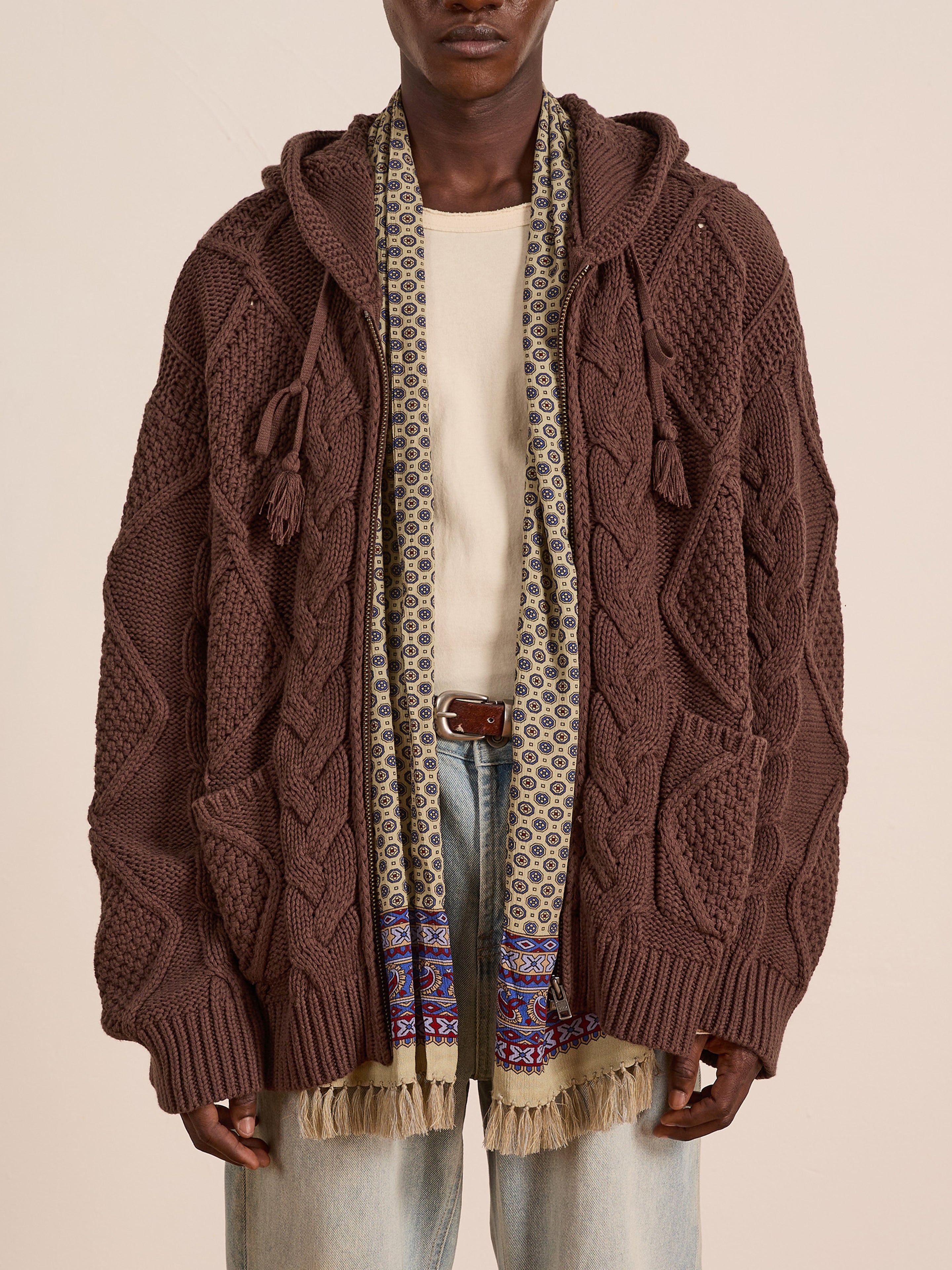 A person wears the FOUND Zip Up Cable Knit Hoodie, a vintage-inspired chunky brown cardigan, over a cream shirt, patterned scarf, and light blue jeans.