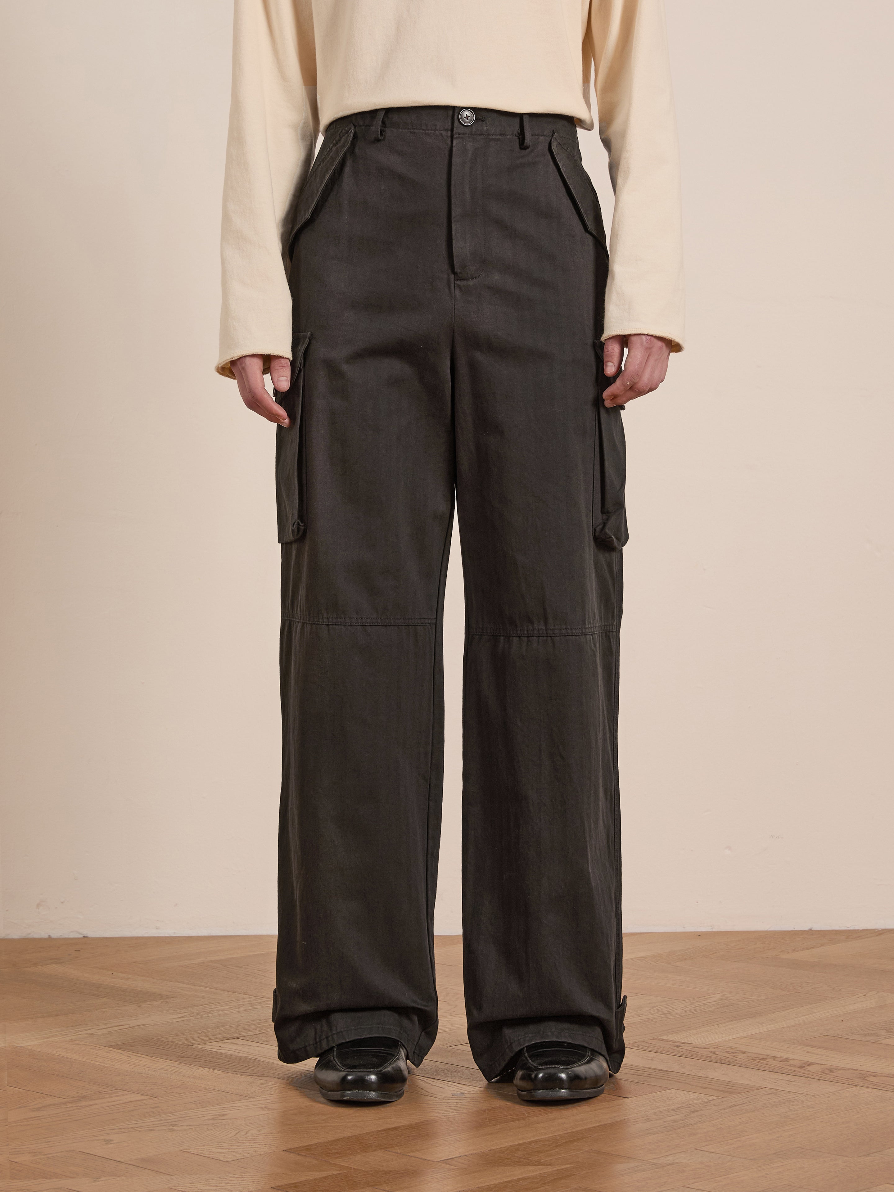 A person in a light beige long-sleeve shirt and FOUND's black Utility Herringbone Cargo Pants stands on a wooden floor indoors, achieving a vintage look with durable cotton twill fabric.