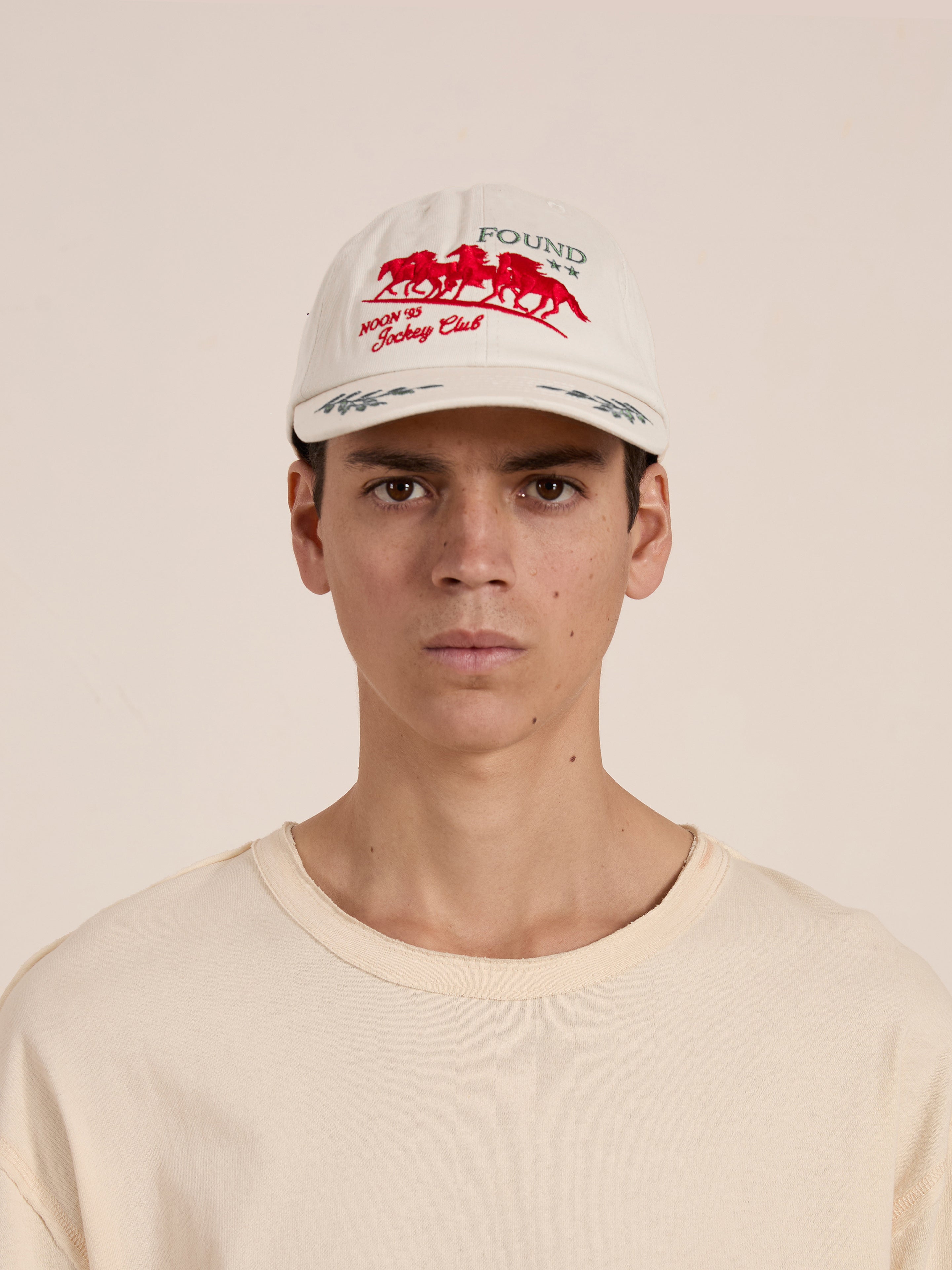 A person wearing a beige shirt and the FOUND Jockey Club Found Cap featuring an embroidered logo of galloping horses with green text stands against a neutral background.