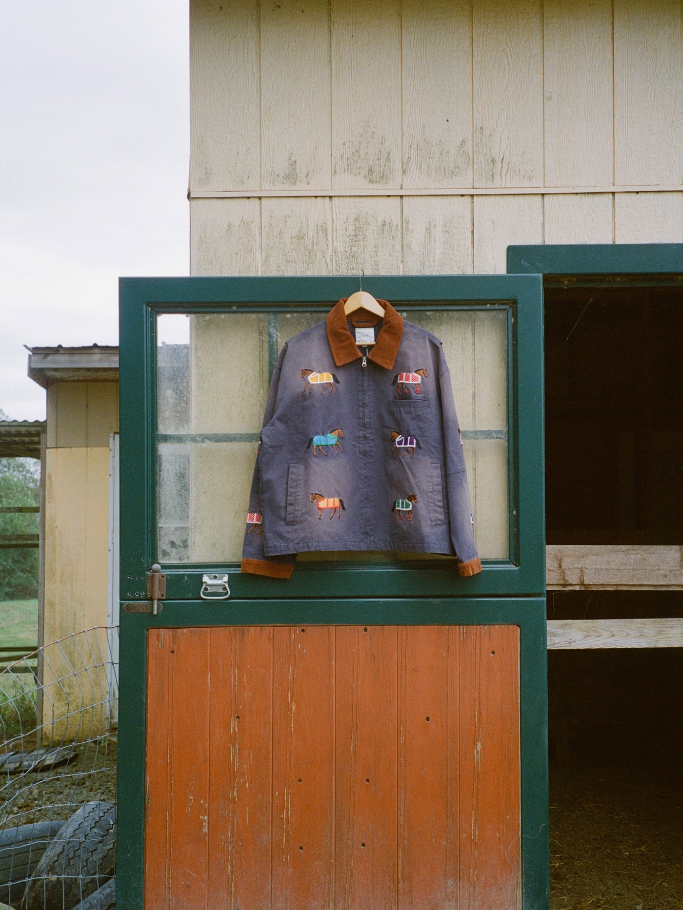 The Found Lariat Horse Embroidered Work Jacket, a unisex navy piece with colorful horse embroidery, hangs on the top half of a Dutch door in front of a barn.