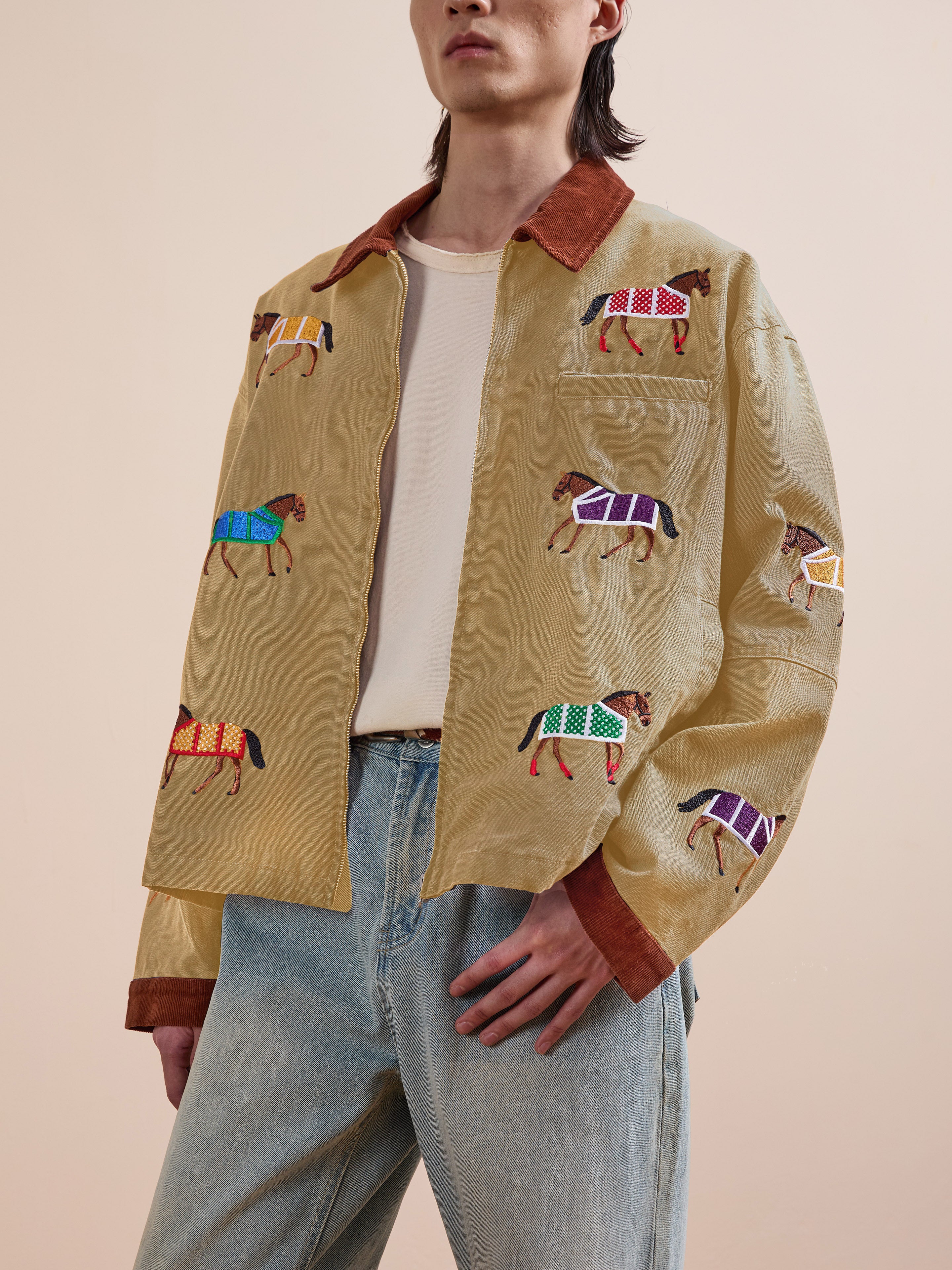 Wearing the Found Lariat Horse Embroidered Work Jacket—tan with multicolored horse designs—over a white shirt and light blue jeans, a person stands against a plain background.