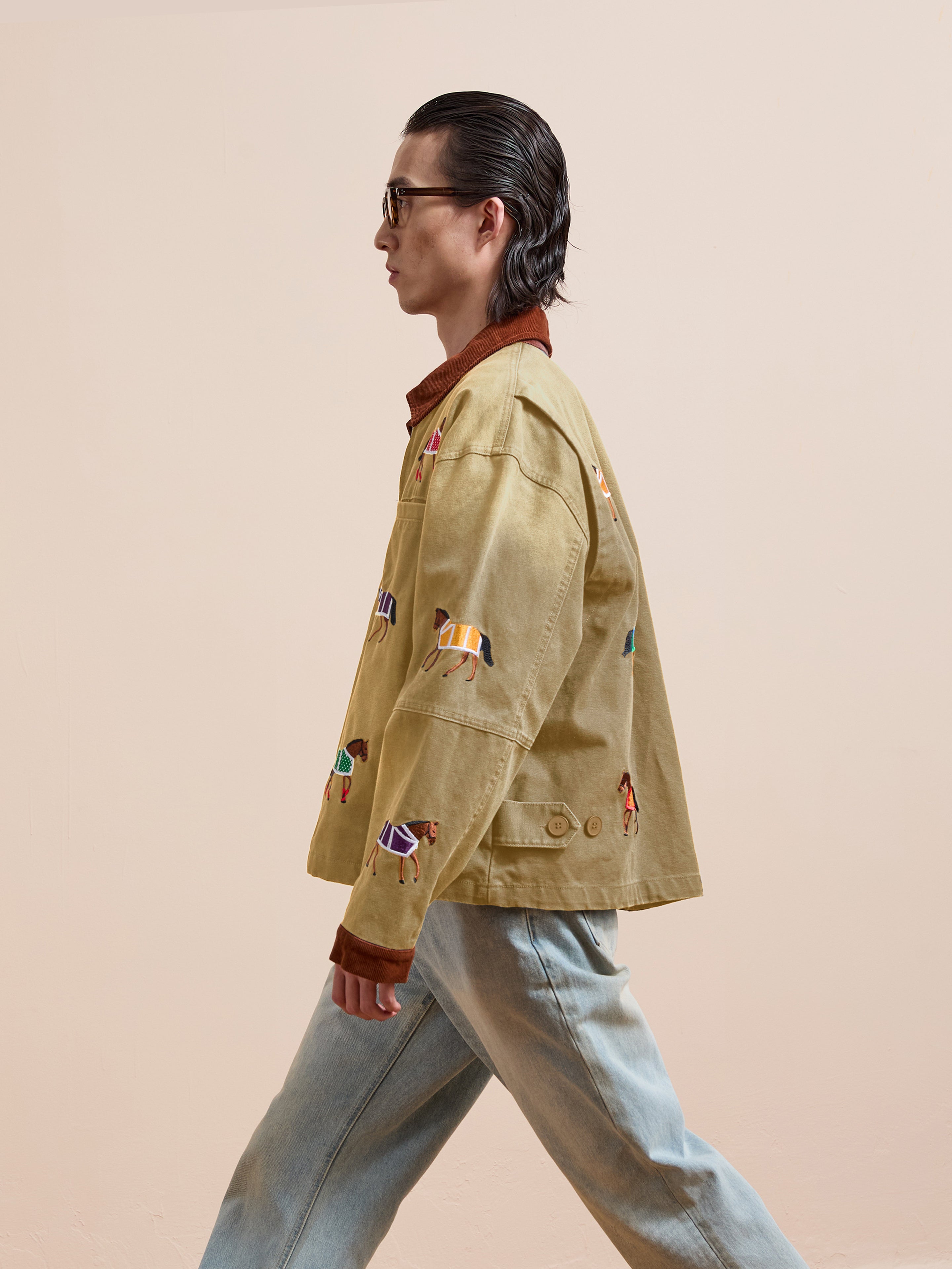 A person with slicked-back hair and glasses wears Found’s Lariat Horse Embroidered Work Jacket in tan and light blue jeans, walking in profile against a plain beige background.