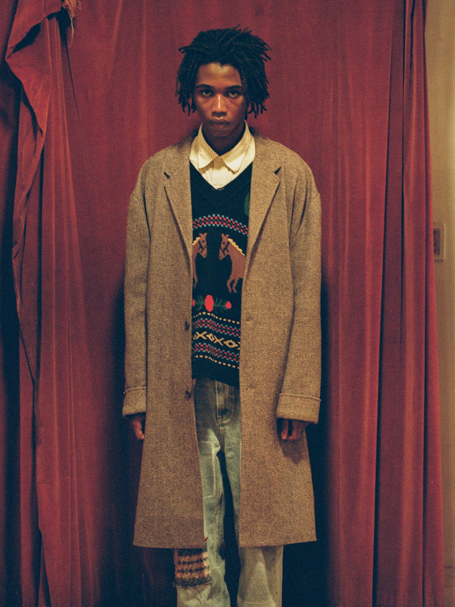 A man wearing the Found Isle Horse Reverie Sweater Knit Vest stands in front of a red curtain.