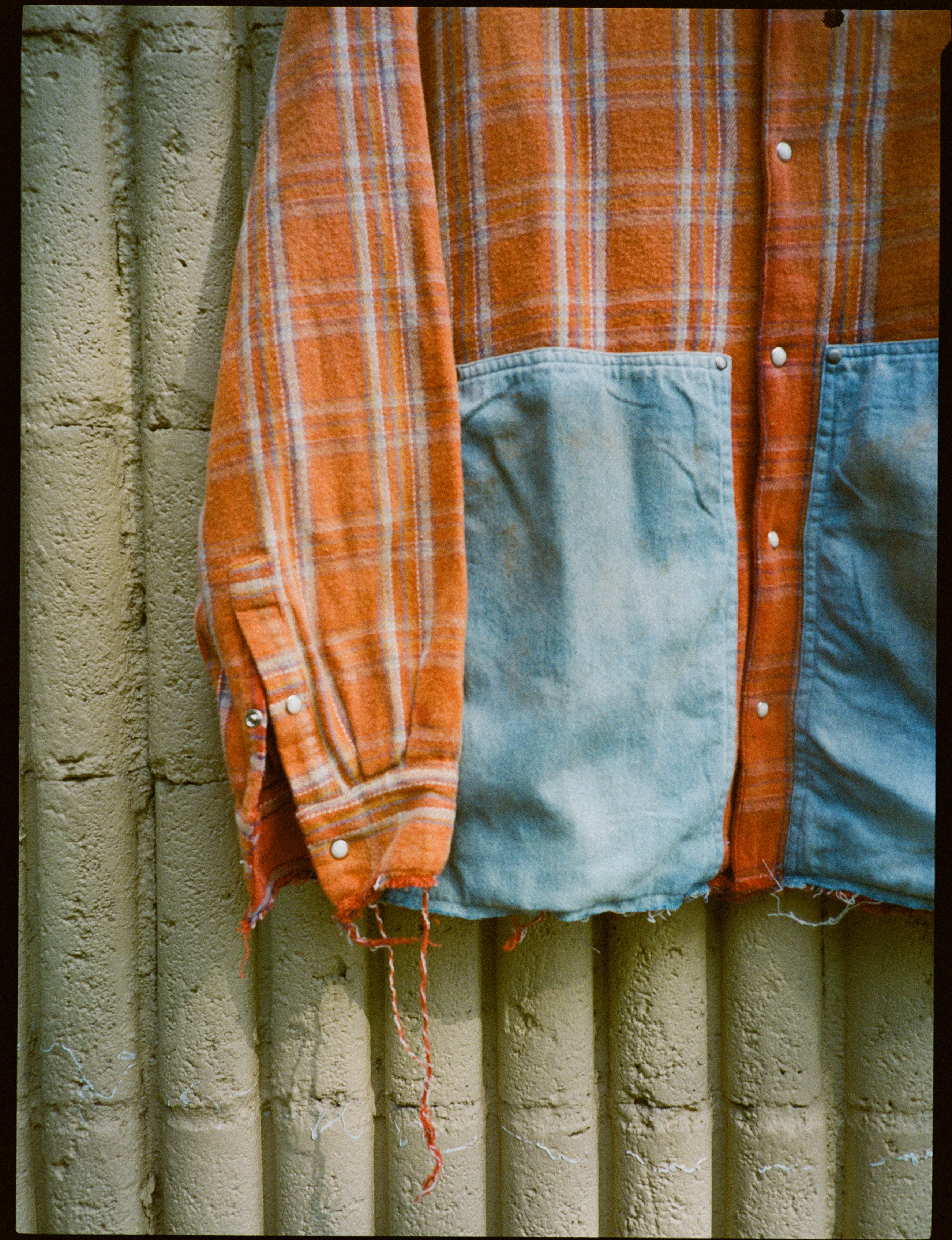 The FOUND Plaid Denim Shirt is a unisex relaxed-fit orange plaid shirt with large blue denim carpenter pockets and frayed edges, shown against a textured beige wall.