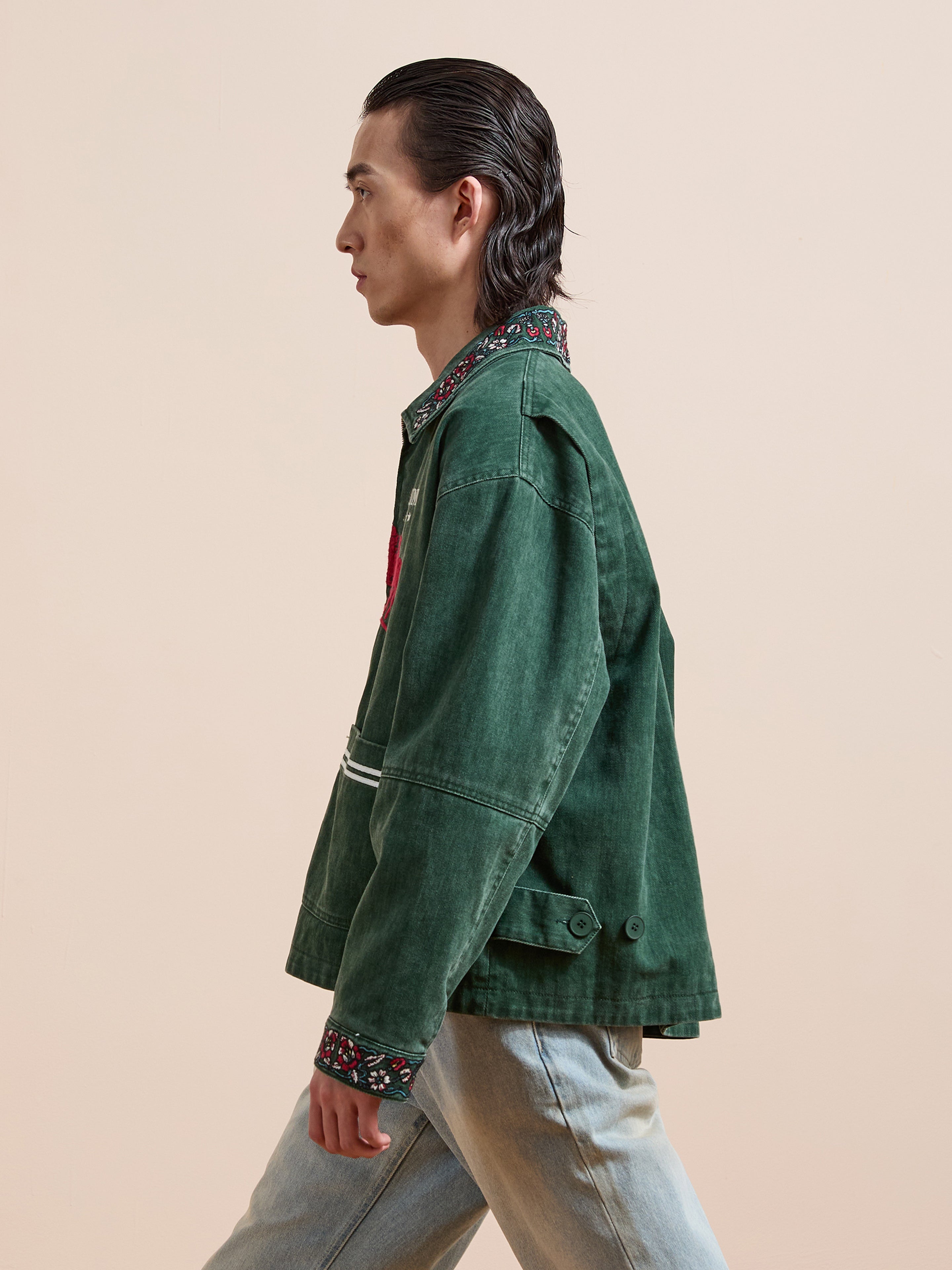 A person with slicked-back hair wears the Found Jockey Club Embroidered Jacket with racing horses and a patterned collar, paired with light blue jeans. The unisex, relaxed-fit look is shown in profile against a plain beige background.