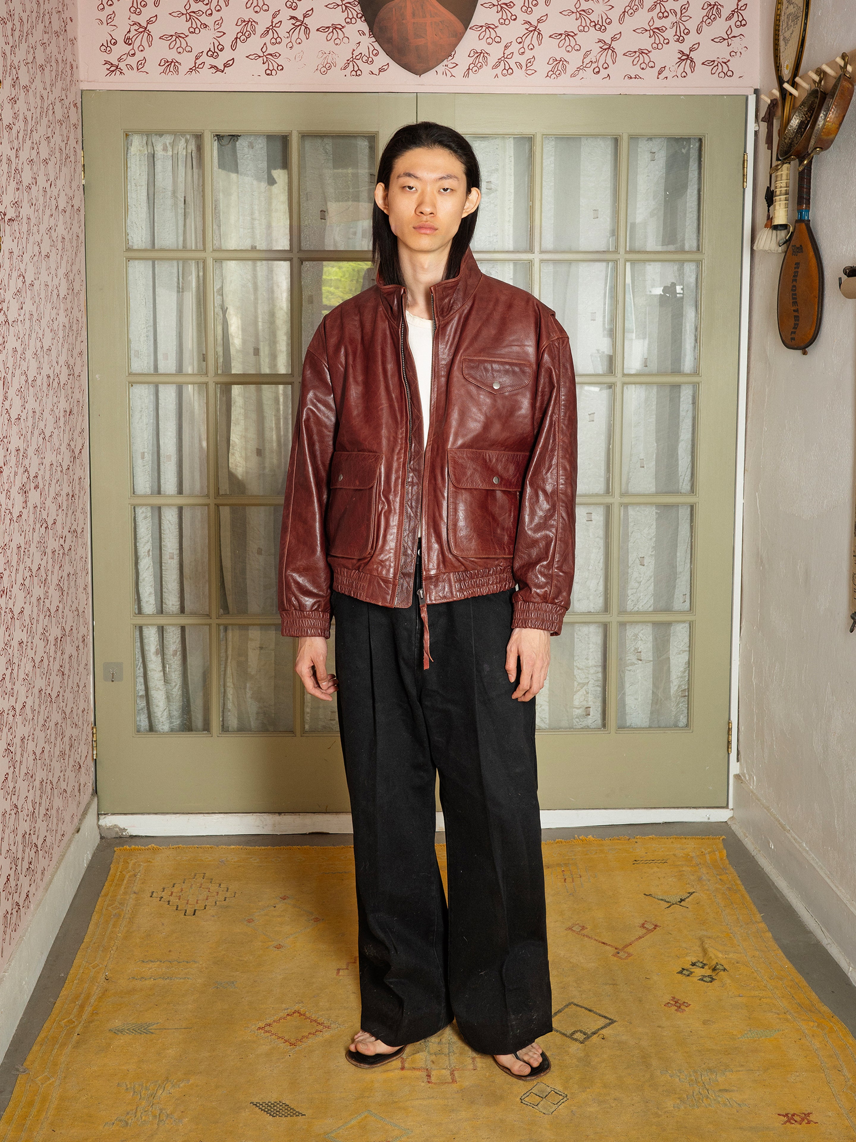 Indoors, a person models the Found Redwood Leather Bomber Jacket—a unisex maroon genuine leather style—paired with a white shirt, black wide-leg pants, and sandals, standing by glass doors and patterned wallpaper.