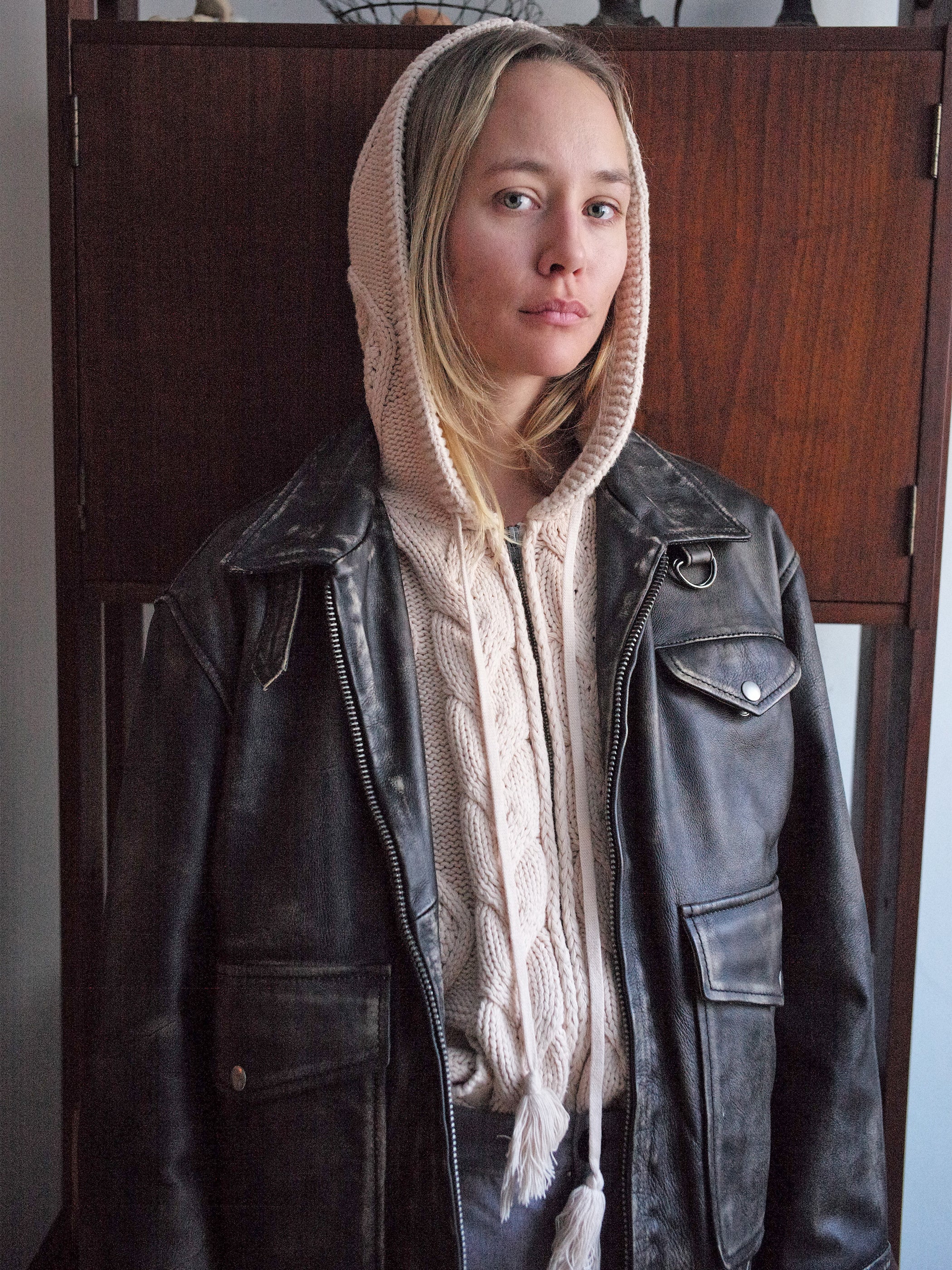 A person stands in front of a wooden cabinet, wearing the Found Distressed Leather Pocket Jacket over a knitted hooded sweater.