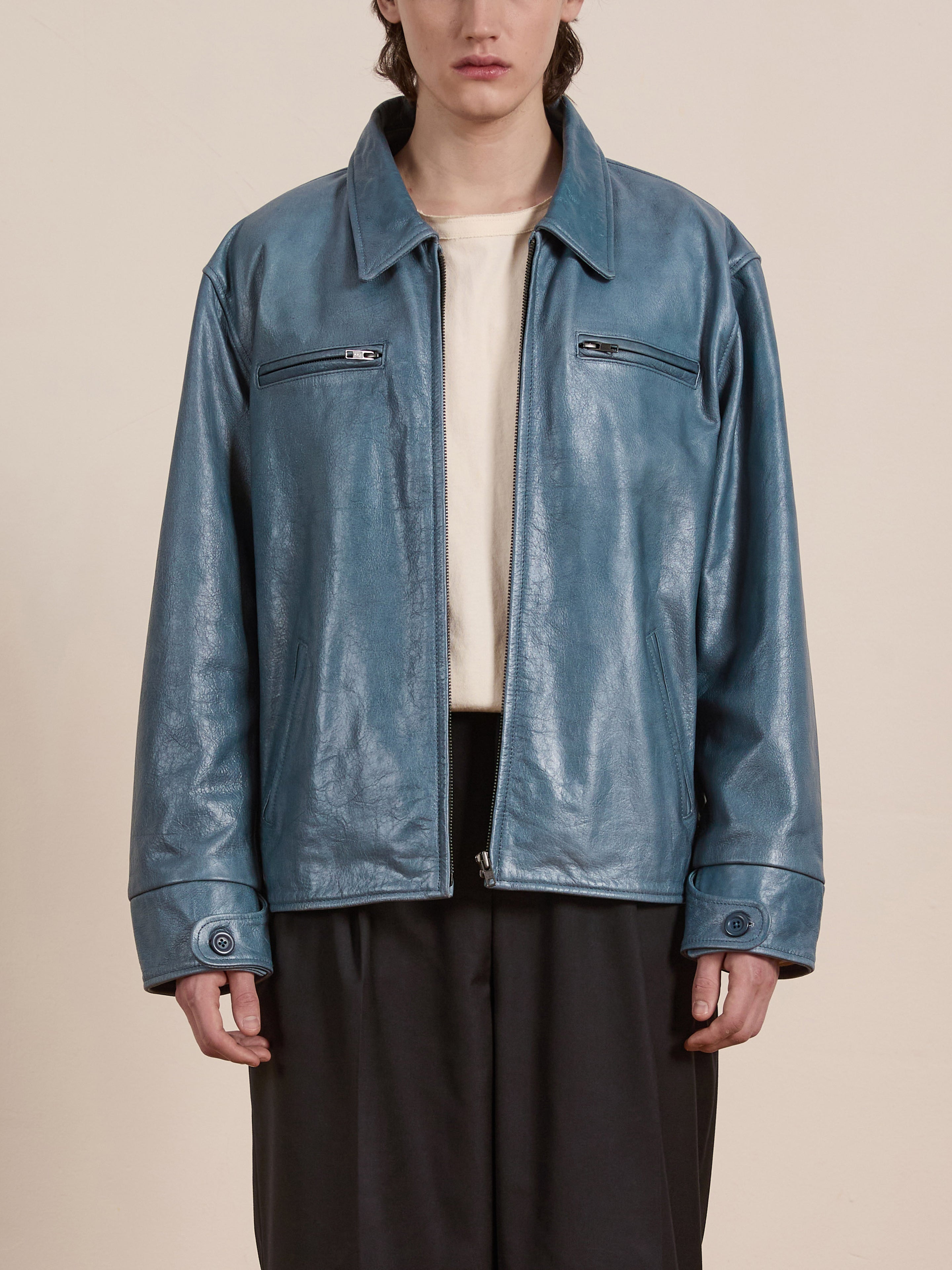 Wearing a Marlboro Sun Faded Leather Racer Jacket by FOUND in teal with a unisex relaxed fit, the person effortlessly drapes it over a white shirt and dark pants against a plain background.
