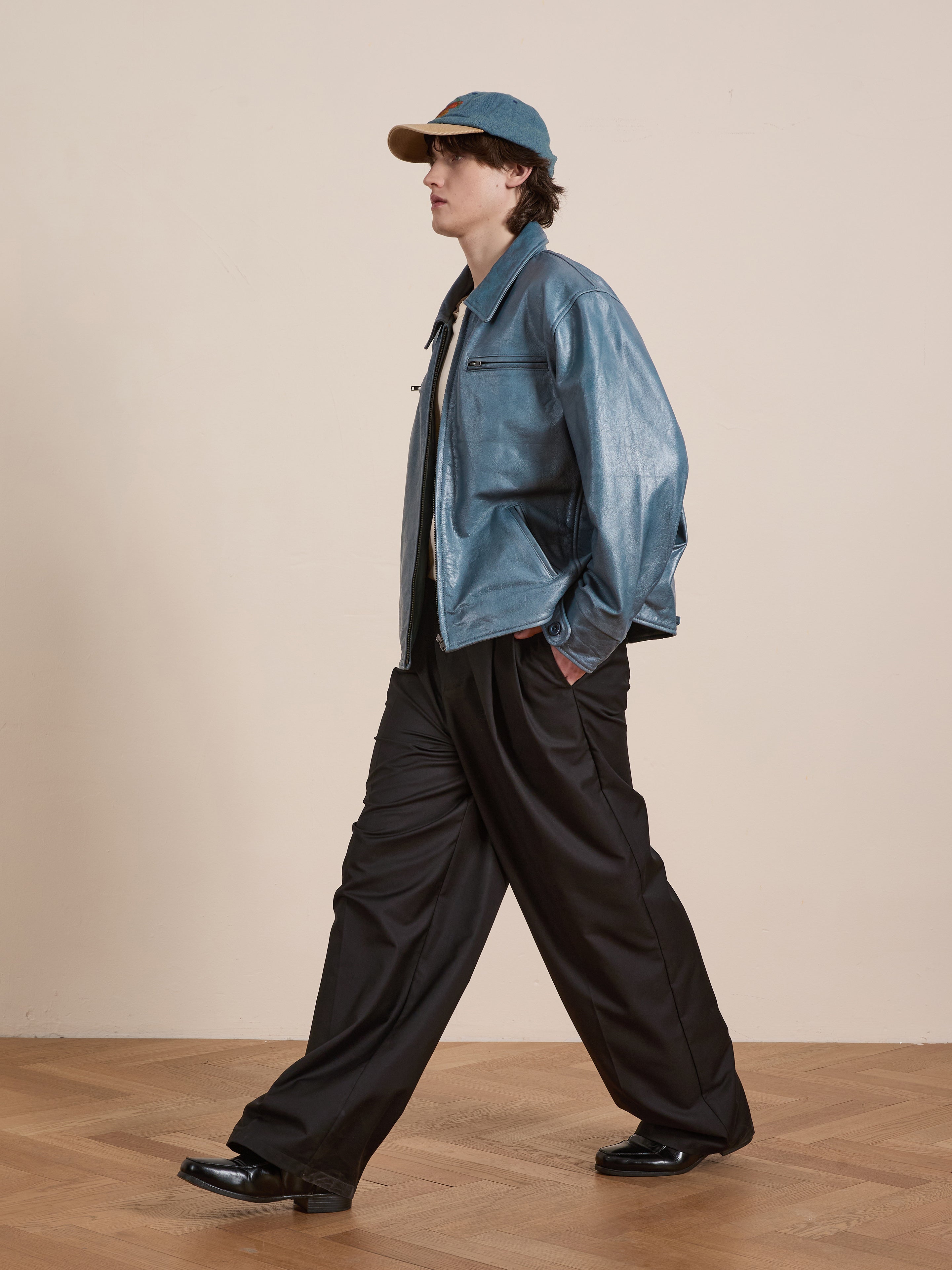 A person wearing a Marlboro Sun Faded Leather Racer Jacket by FOUND, black wide-leg pants, and a blue cap walks on a wooden floor against a light background.