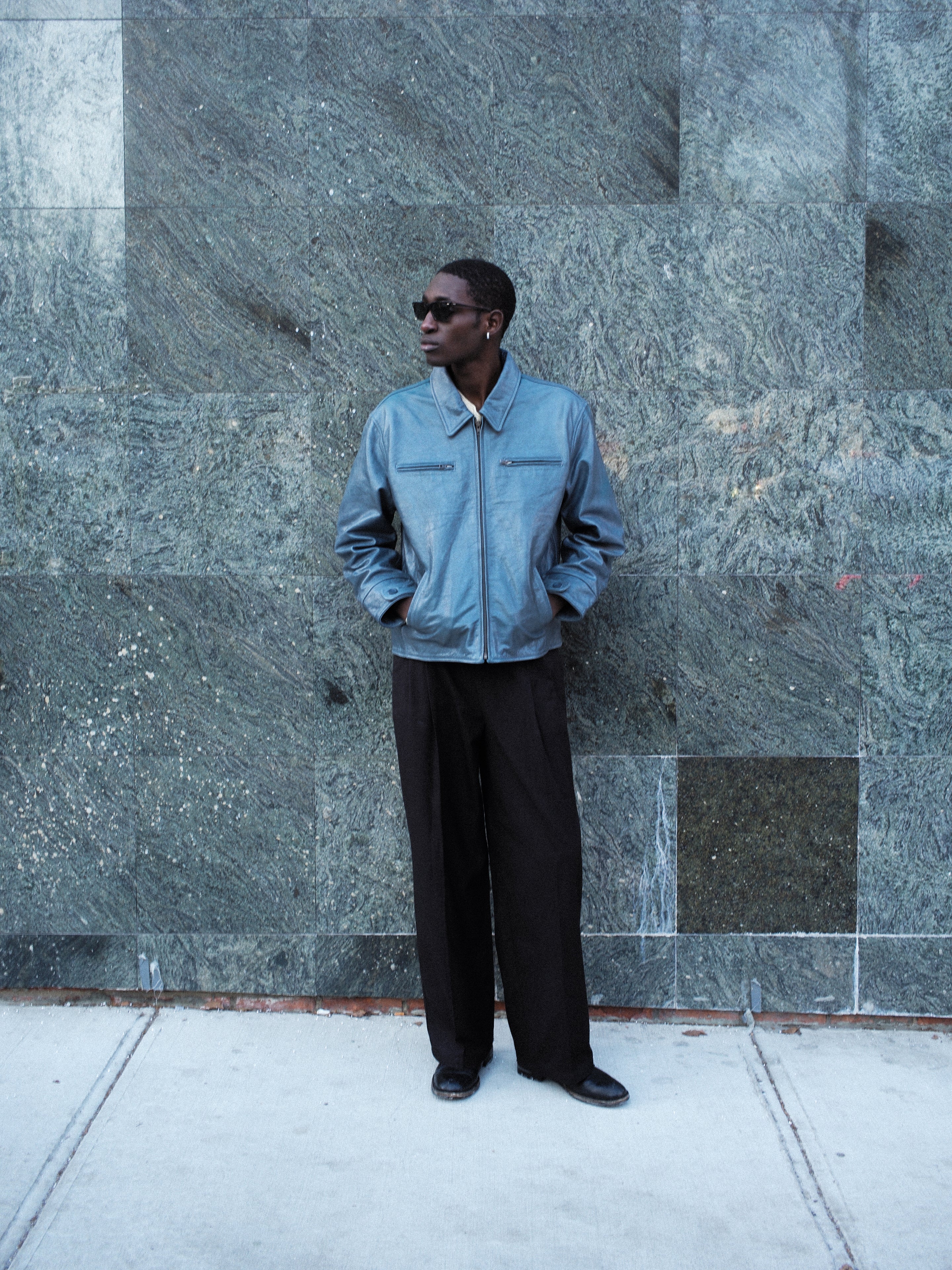 Wearing FOUND's unisex Marlboro Sun Faded Leather Racer Jacket and black pants, a person in sunglasses stands against a textured stone wall, gazing to the side.