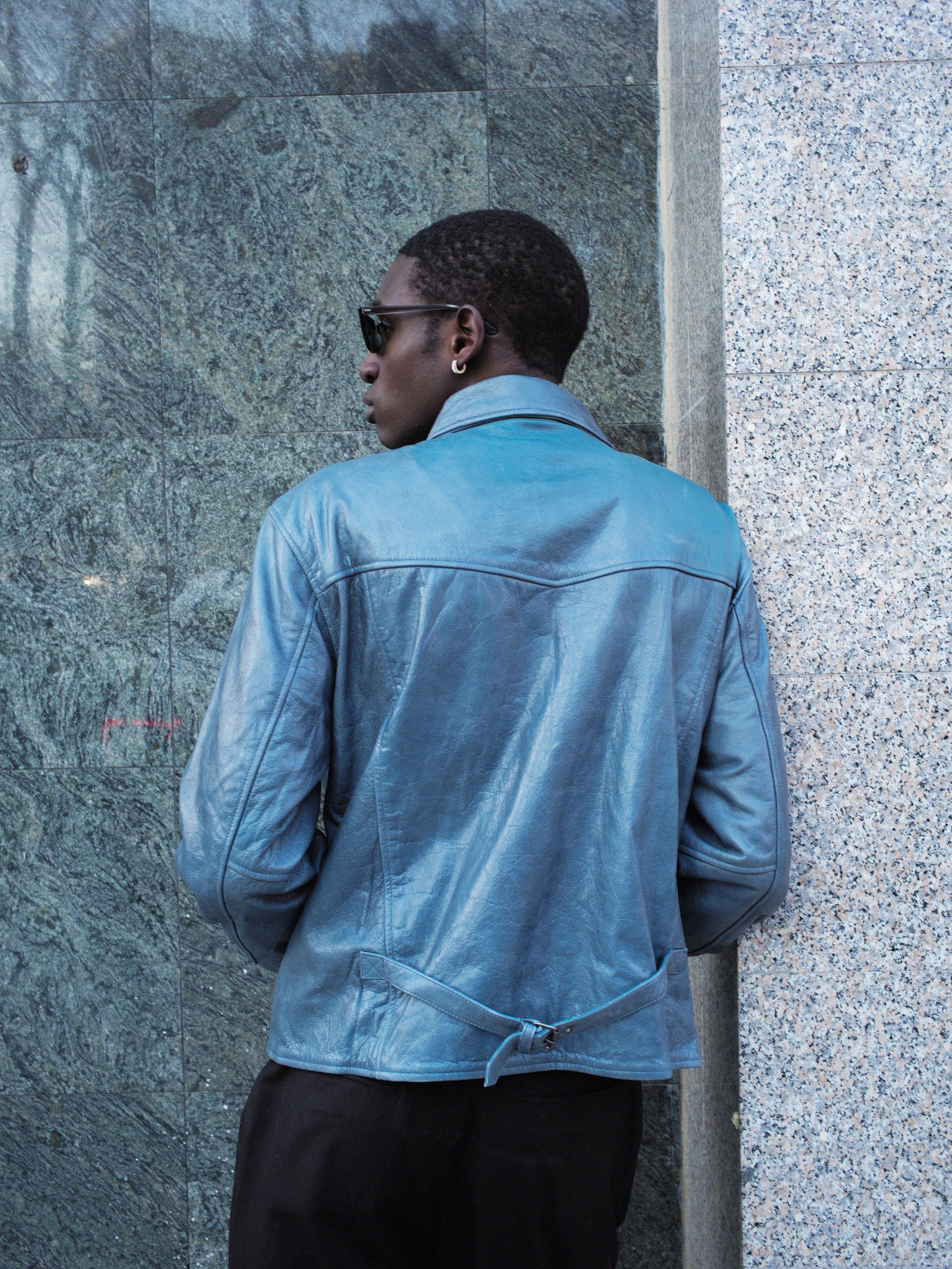 A person in FOUND's Marlboro Sun Faded Leather Racer Jacket, wearing sunglasses, stands with their back to the camera against a stone wall.