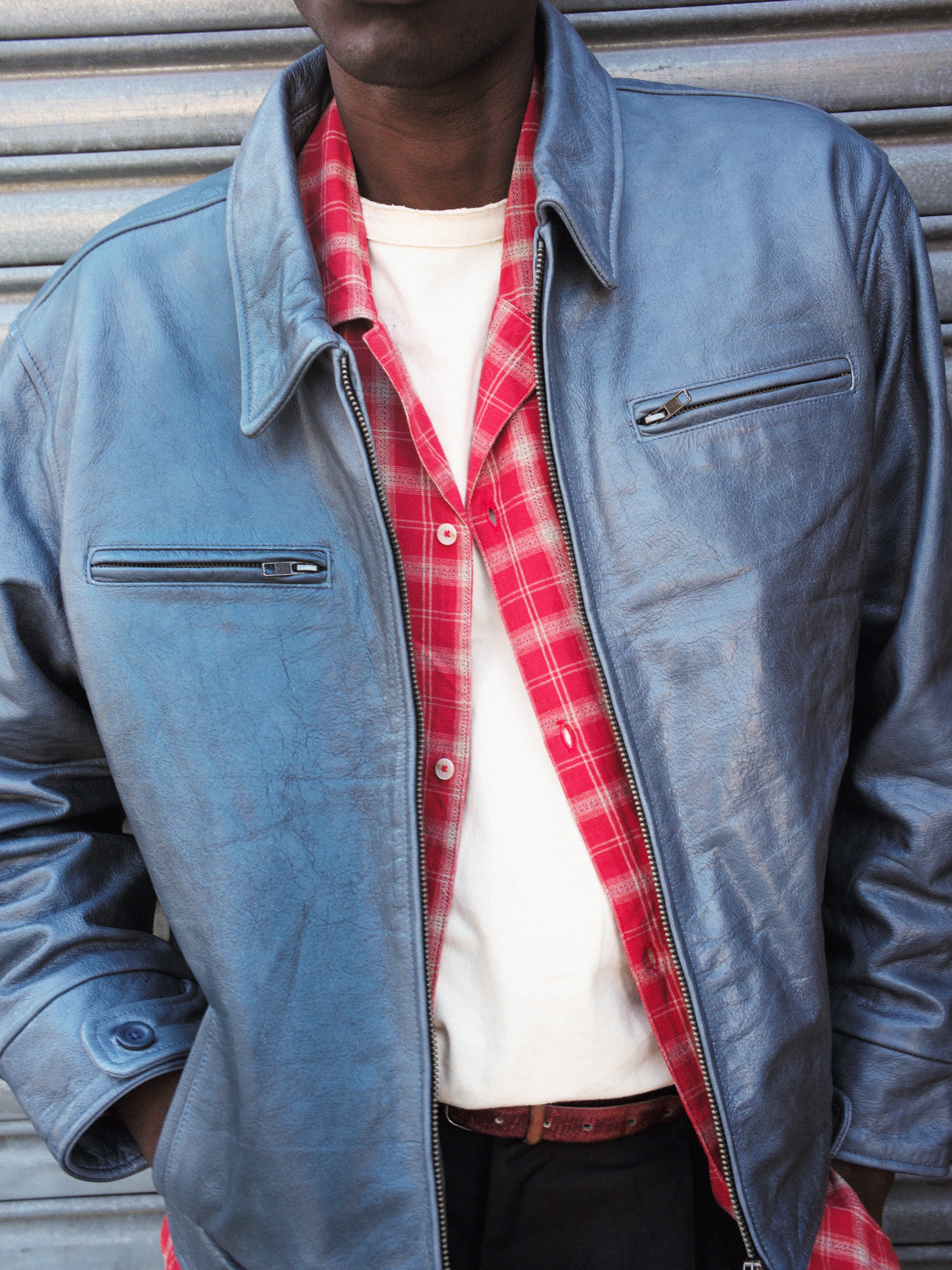 Dressed in a Marlboro Sun Faded Leather Racer Jacket by FOUND over a red plaid shirt and white T-shirt, a person stands confidently against a corrugated metal backdrop.