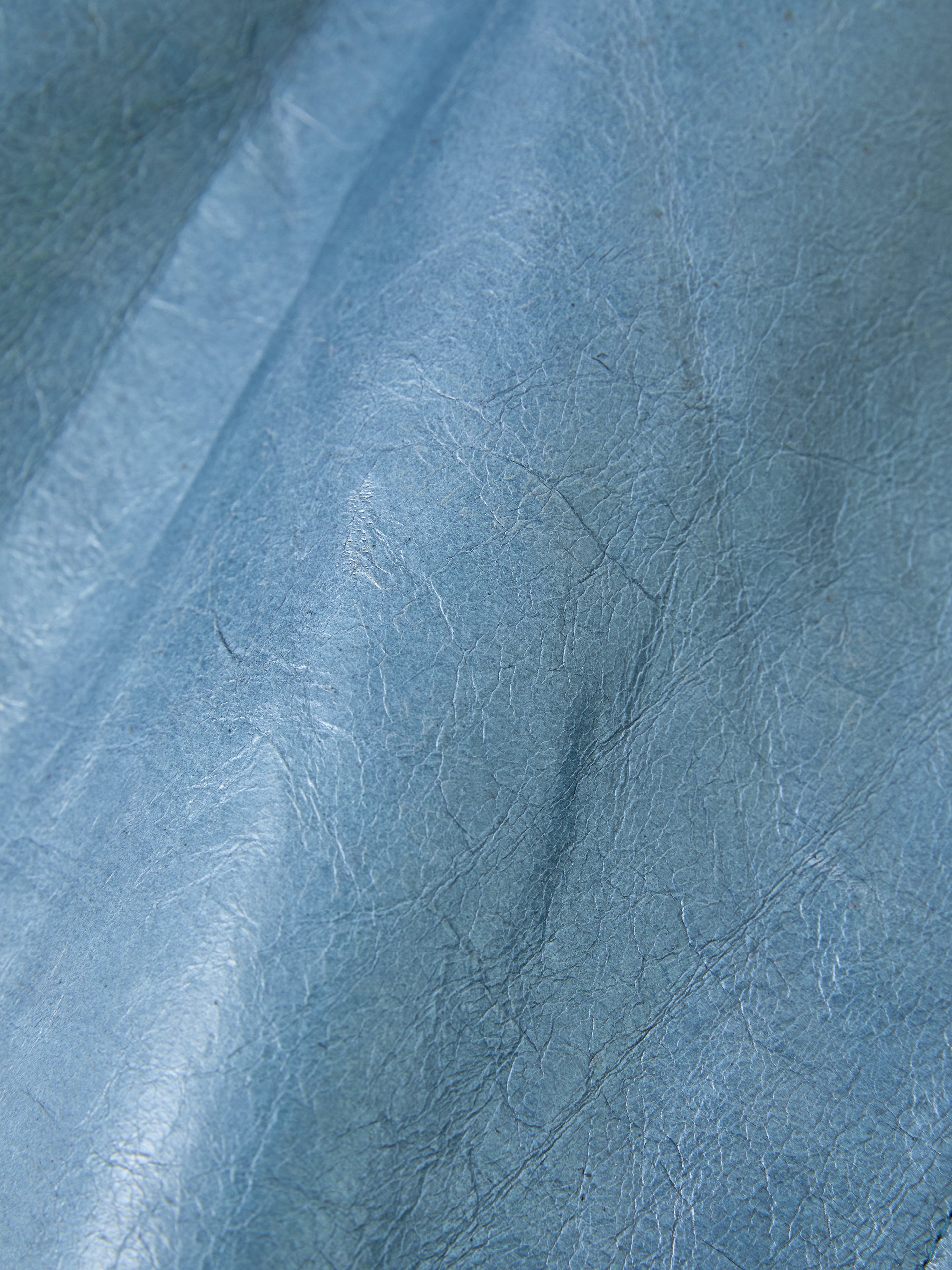 A close-up of the Marlboro Sun Faded Leather Racer Jacket by FOUND showcases its textured, wrinkled blue leather with visible creases and a subtle sheen, exuding an earthy charm.