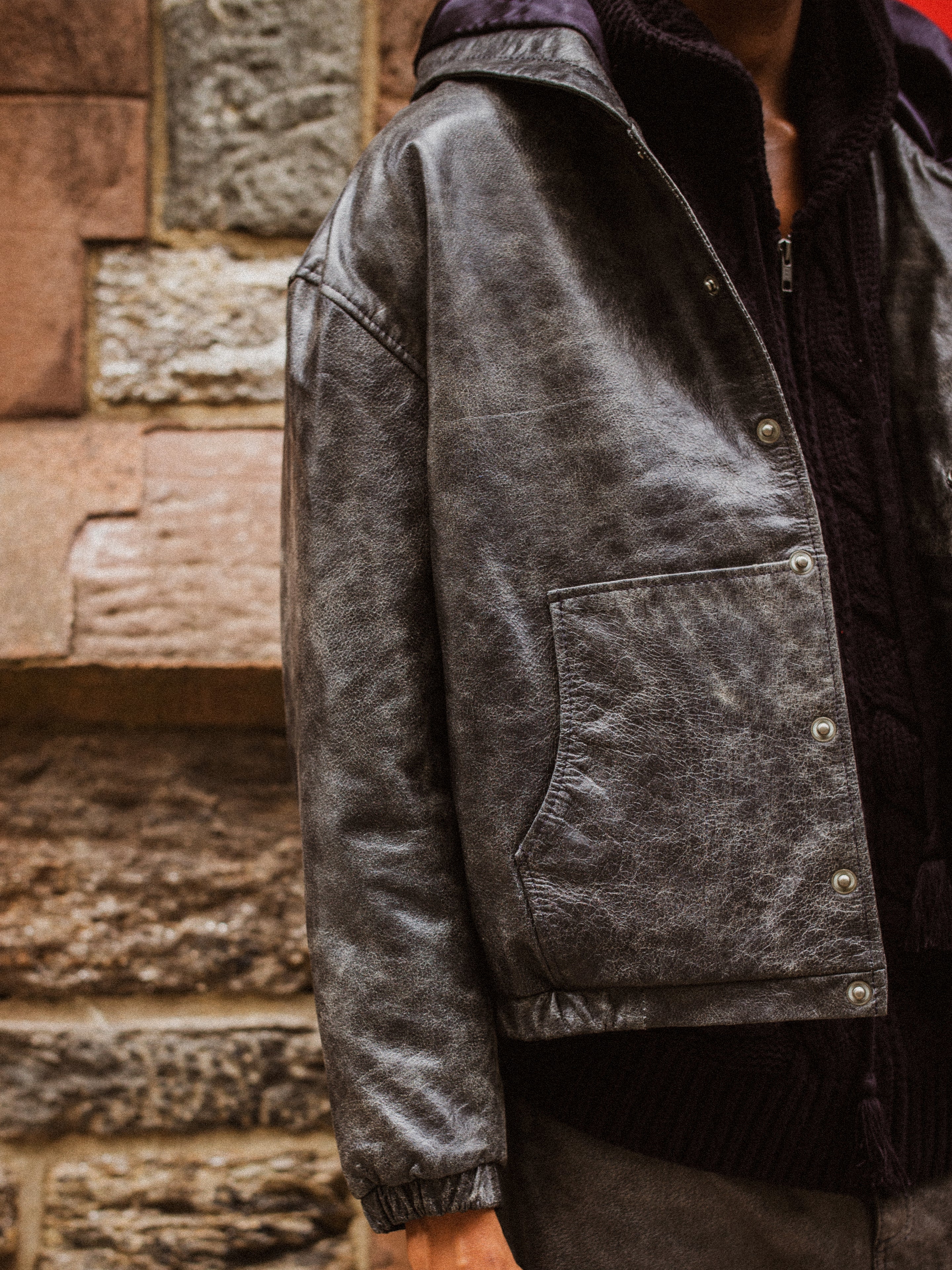 A person wearing Found's dark, lightweight Distressed Genuine Leather Hoodie with a side pocket stands in front of a stone wall. Only part of the person's upper body is visible.