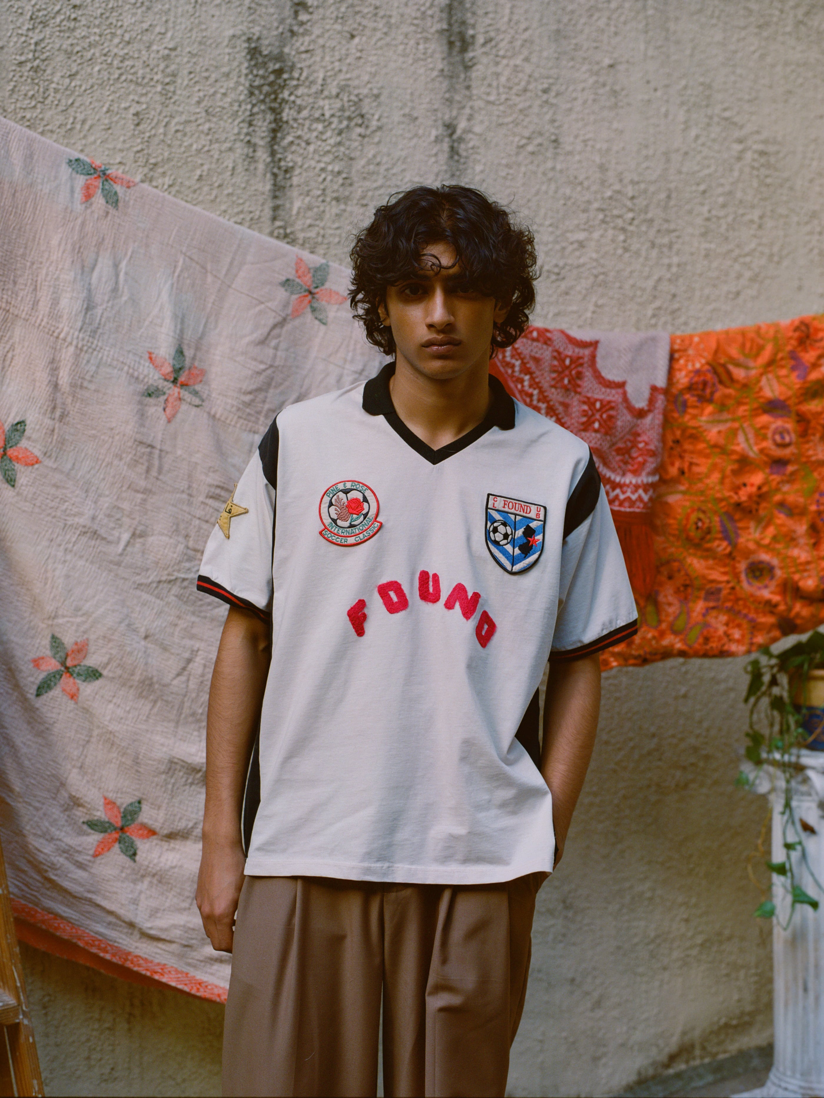 A person with curly hair wearing a Union Soccer Jersey by FOUND stands in front of patterned fabric draped over a line, adding a contemporary edge to the scene.