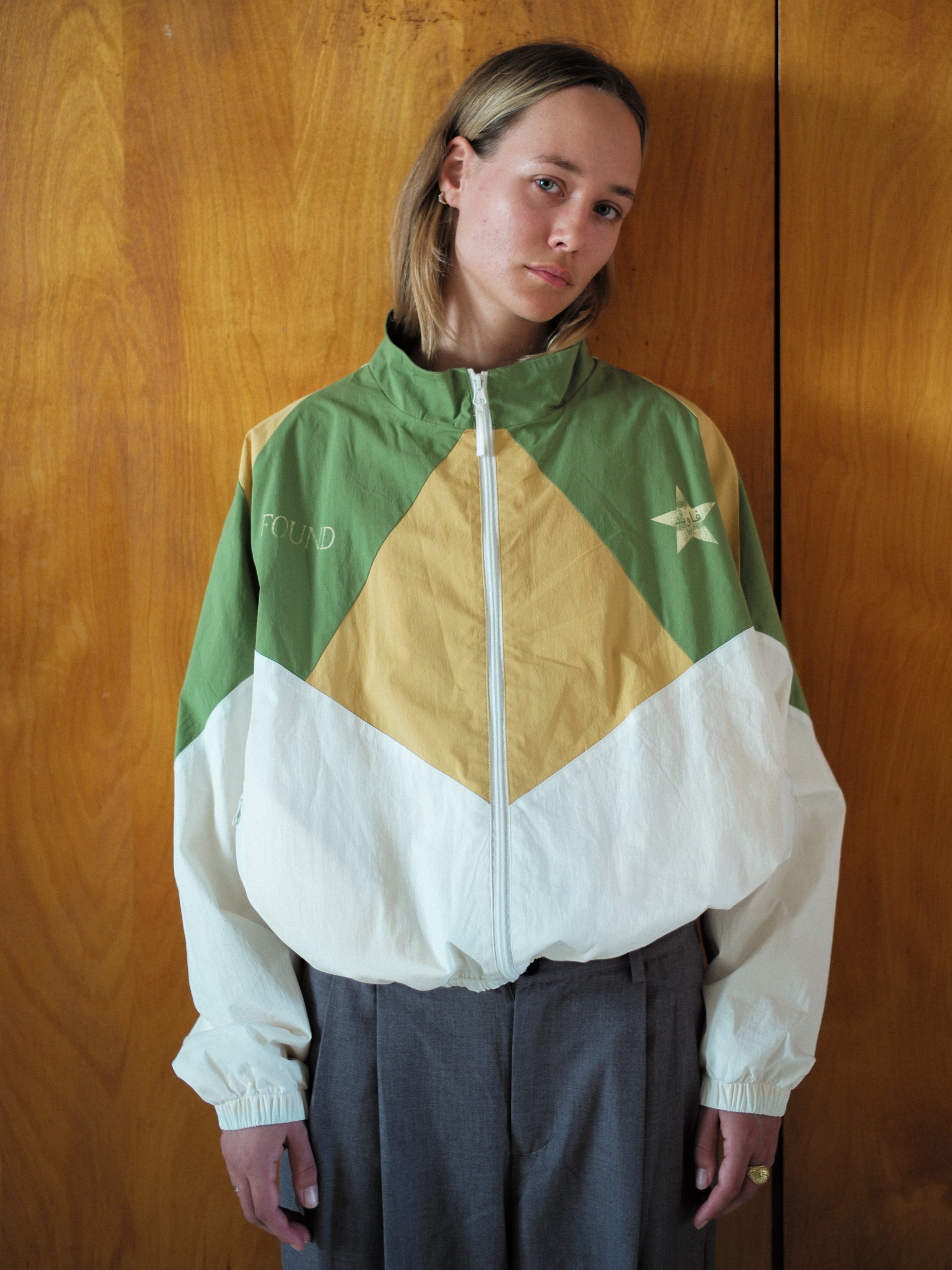Against a wooden backdrop, the individual wears the Legacy Cricket Track Jacket by FOUND in green, yellow, and white with a star and "FOUND" on it. Their straight hair frames a neutral expression, effortlessly combining retro international tracksuits style with timeless appeal.