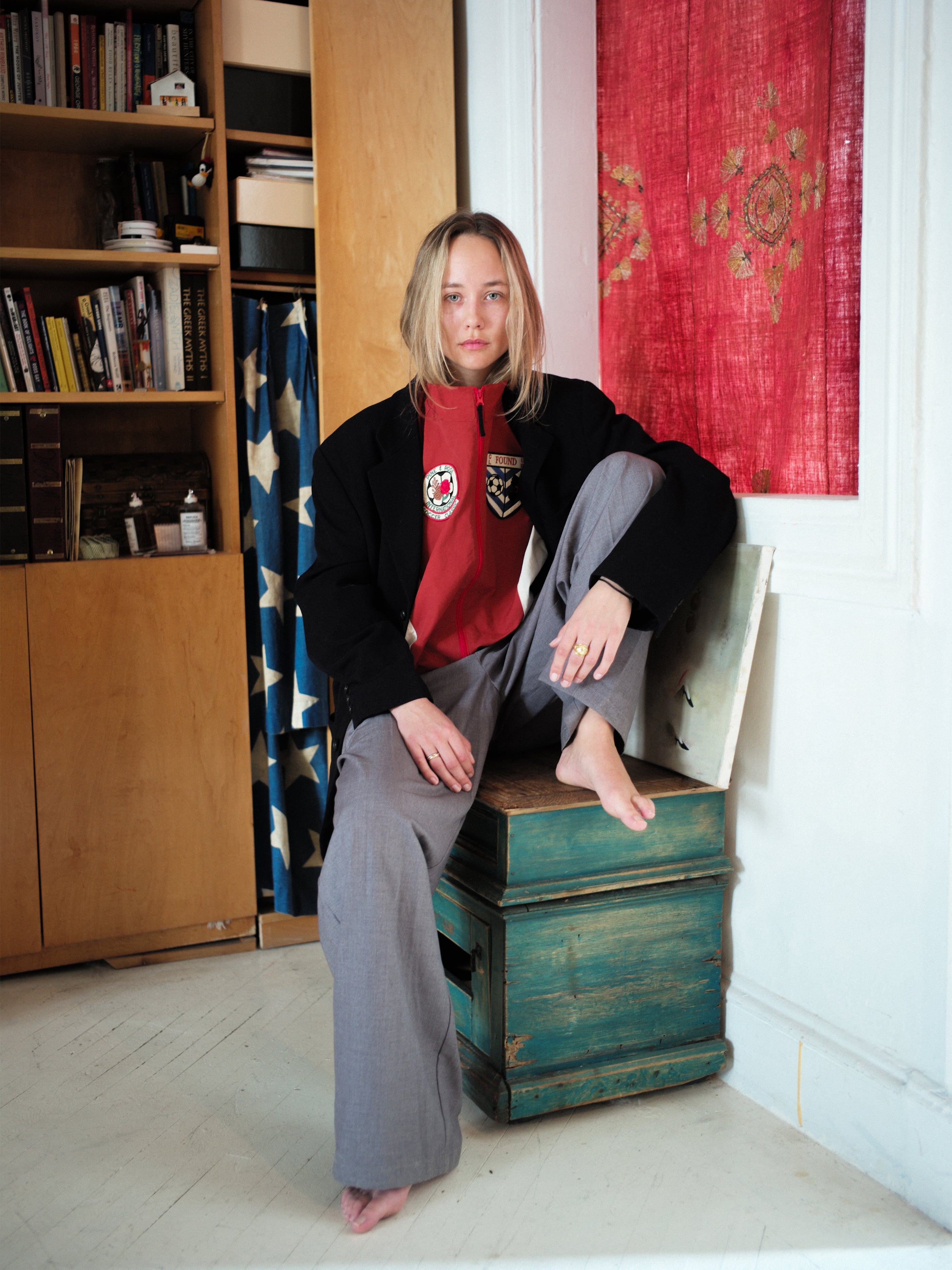 A person in a Pine and Rose Track Jacket by FOUND, paired with a red shirt, sits barefoot on a teal wooden box, one leg crossed. Bookshelves and a red curtain in the background enhance the casual sportswear ambiance.
