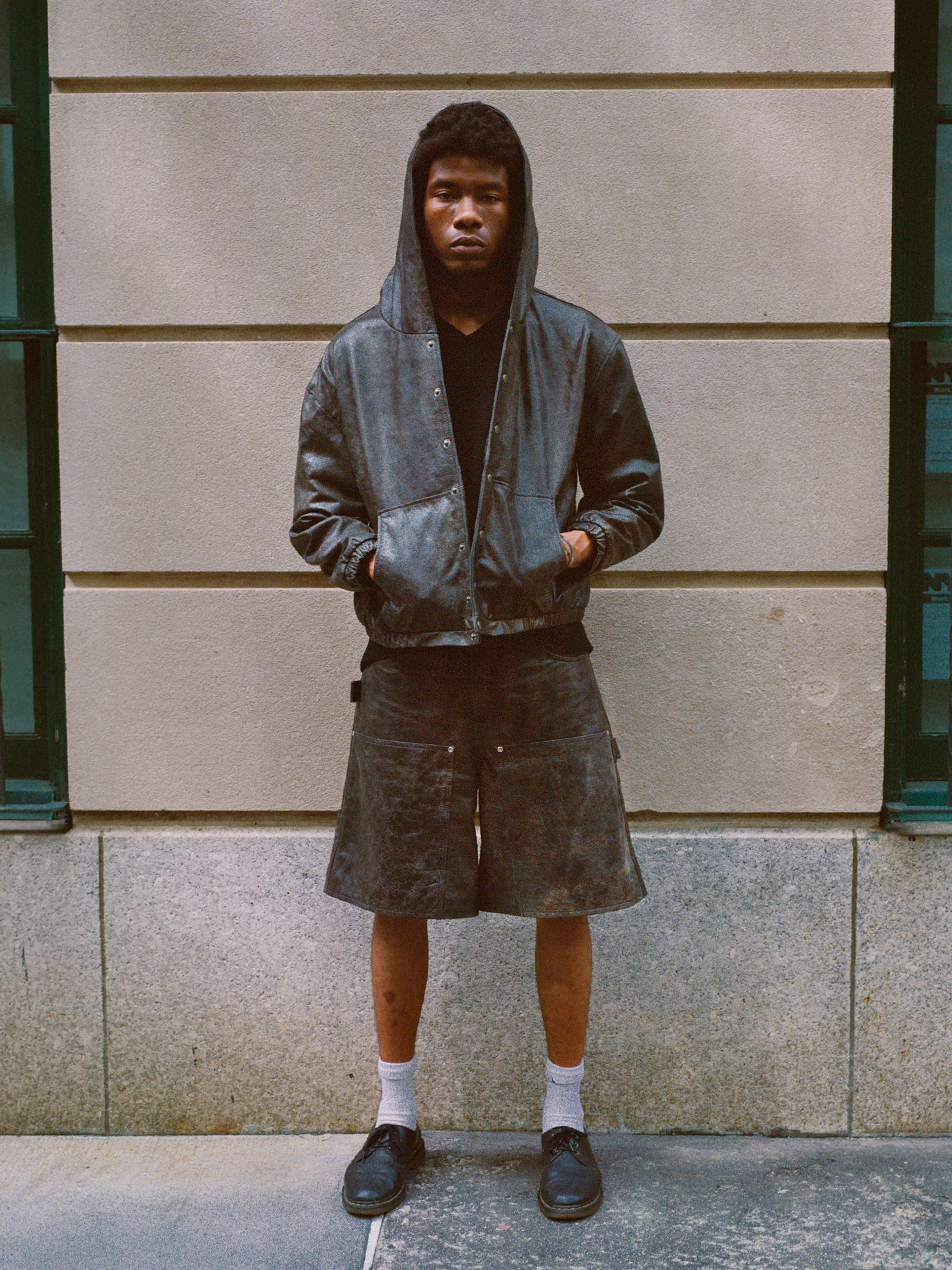 A person stands in front of a neutral-colored wall, wearing the Distressed Genuine Leather Hoodie by Found along with shorts, socks, and shoes. Their hands are in the hoodie pockets and they have a neutral expression on their face.
