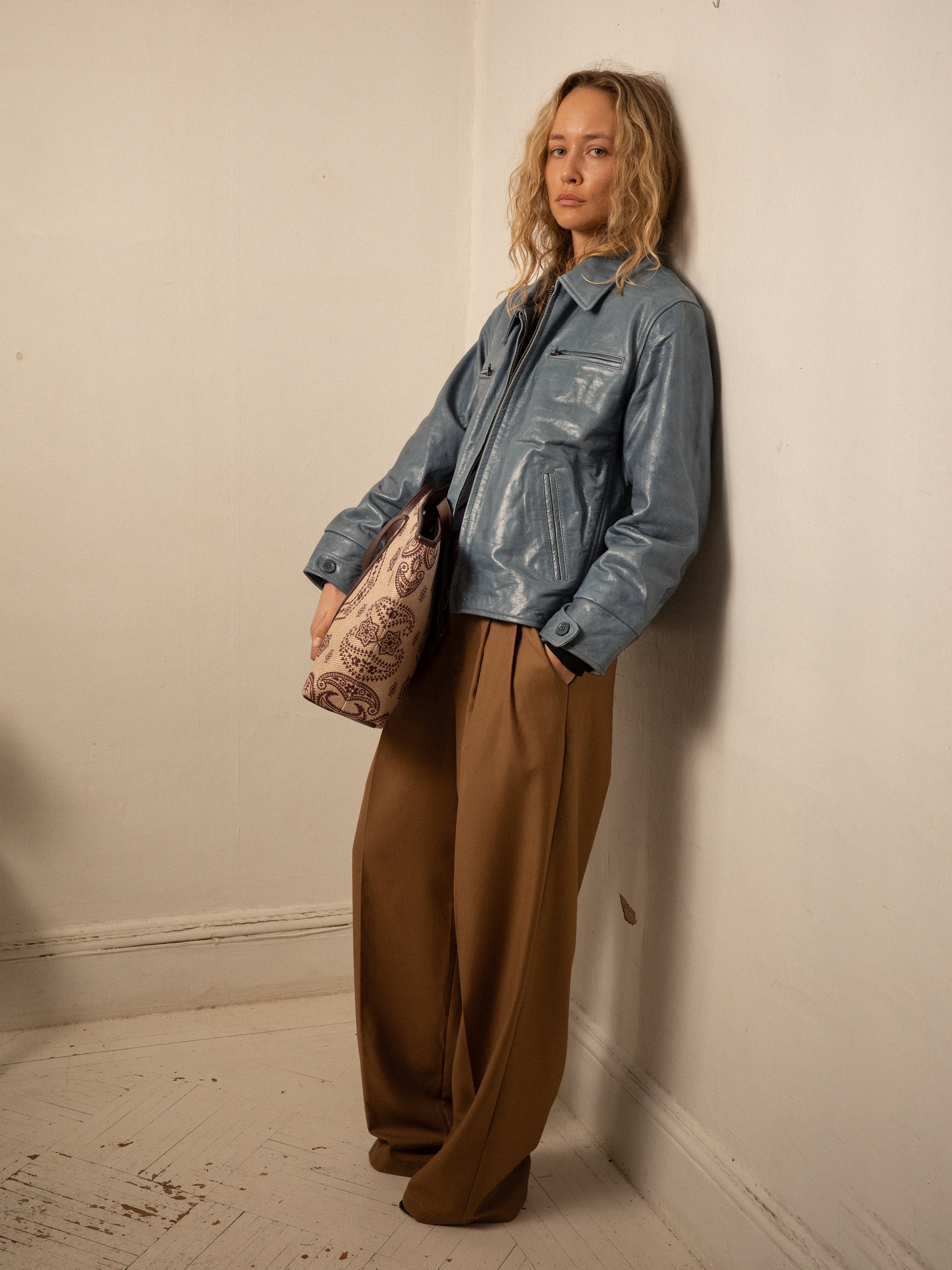A person with curly hair wears a Marlboro Sun Faded Leather Racer Jacket from FOUND paired with tan pants, while casually holding a patterned bag. The unisex relaxed fit suits their style as they stand against the wall on a wooden floor.