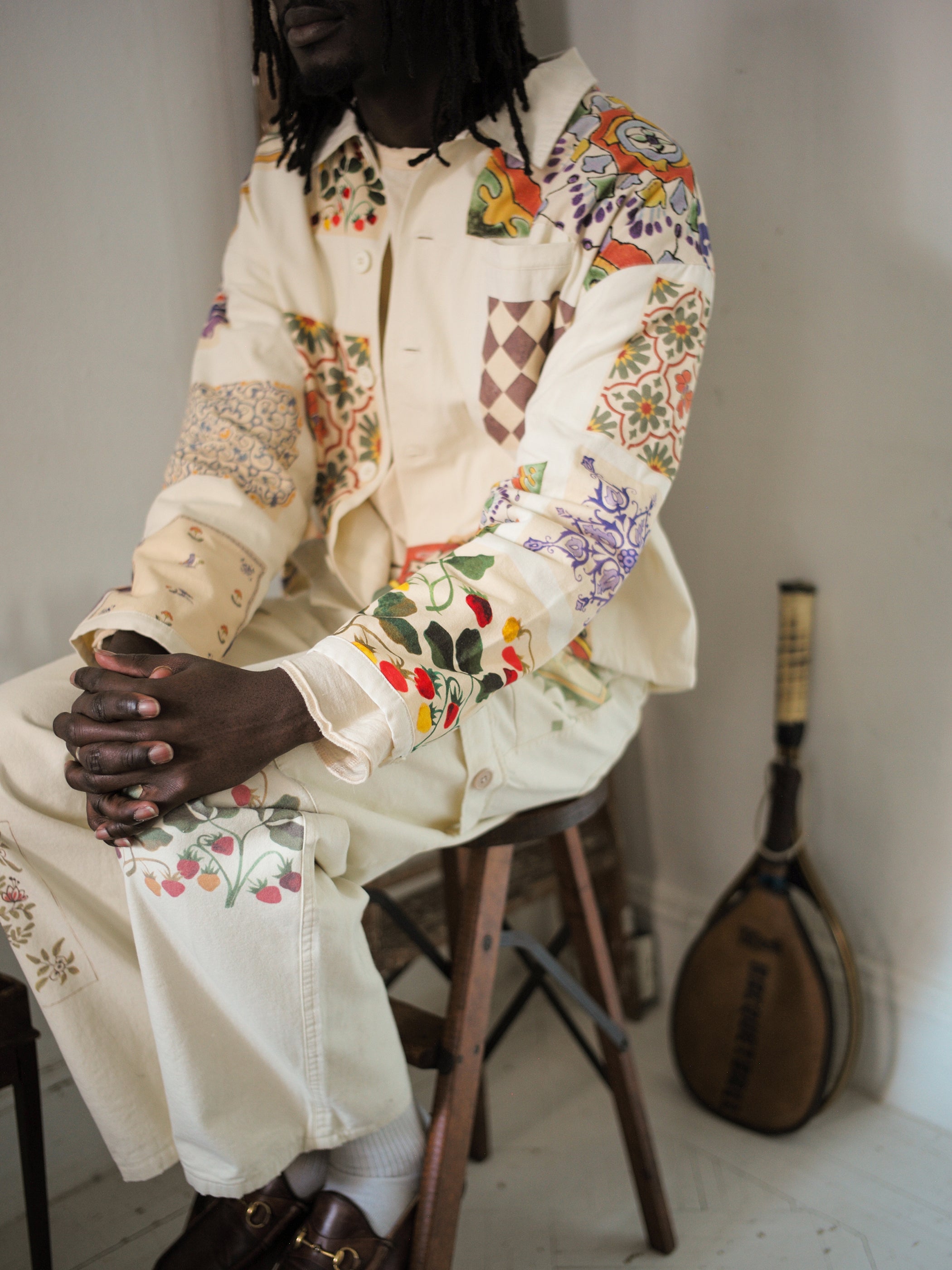 Seated on a stool, a person dons a pair of Mosaic Pants by FOUND in cream, featuring vibrant Indo-Aryan patterns. Behind them, a stringed instrument leans against the wall, enhancing the eclectic and artistic vibe of this unisex attire.