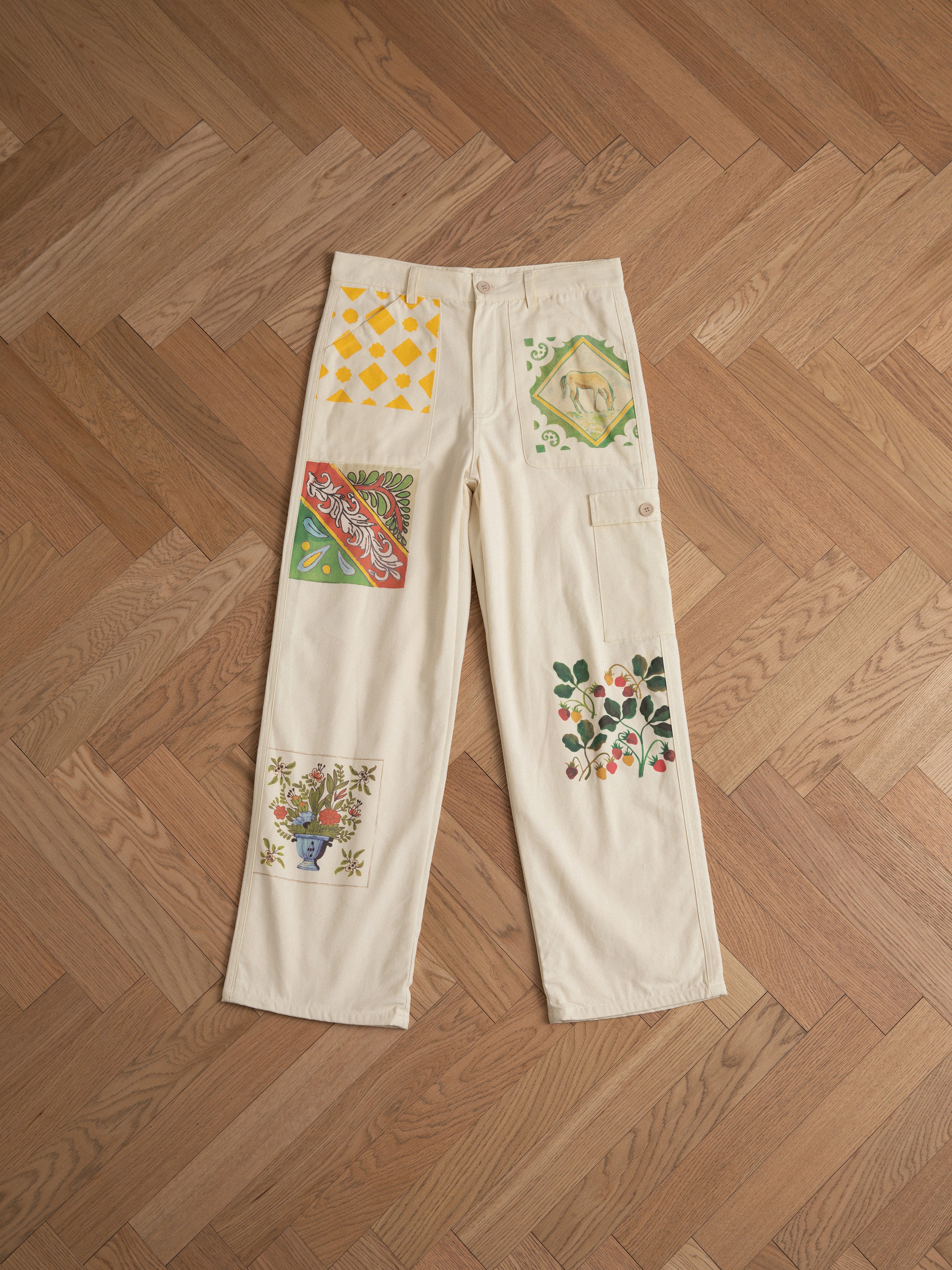 FOUND's Mosaic Pants in cream showcase vibrant abstract designs with floral and Indo-Aryan patterns, laid flat on a herringbone wood floor. Unisex and ideal for a bold fashion statement.