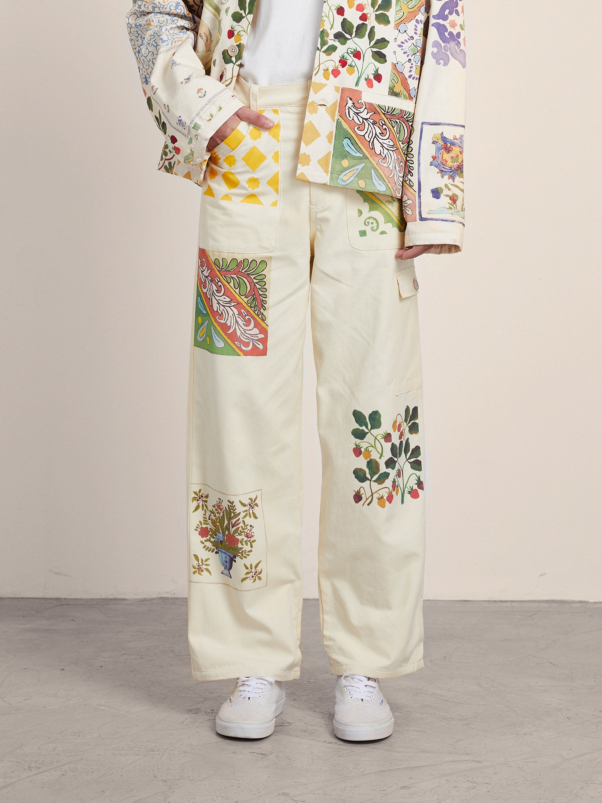A person in FOUND's Mosaic Pants and an Indo-Aryan patterned jacket with floral and geometric designs stands gracefully on a gray floor, captivating with the vibrant unisex ensemble.