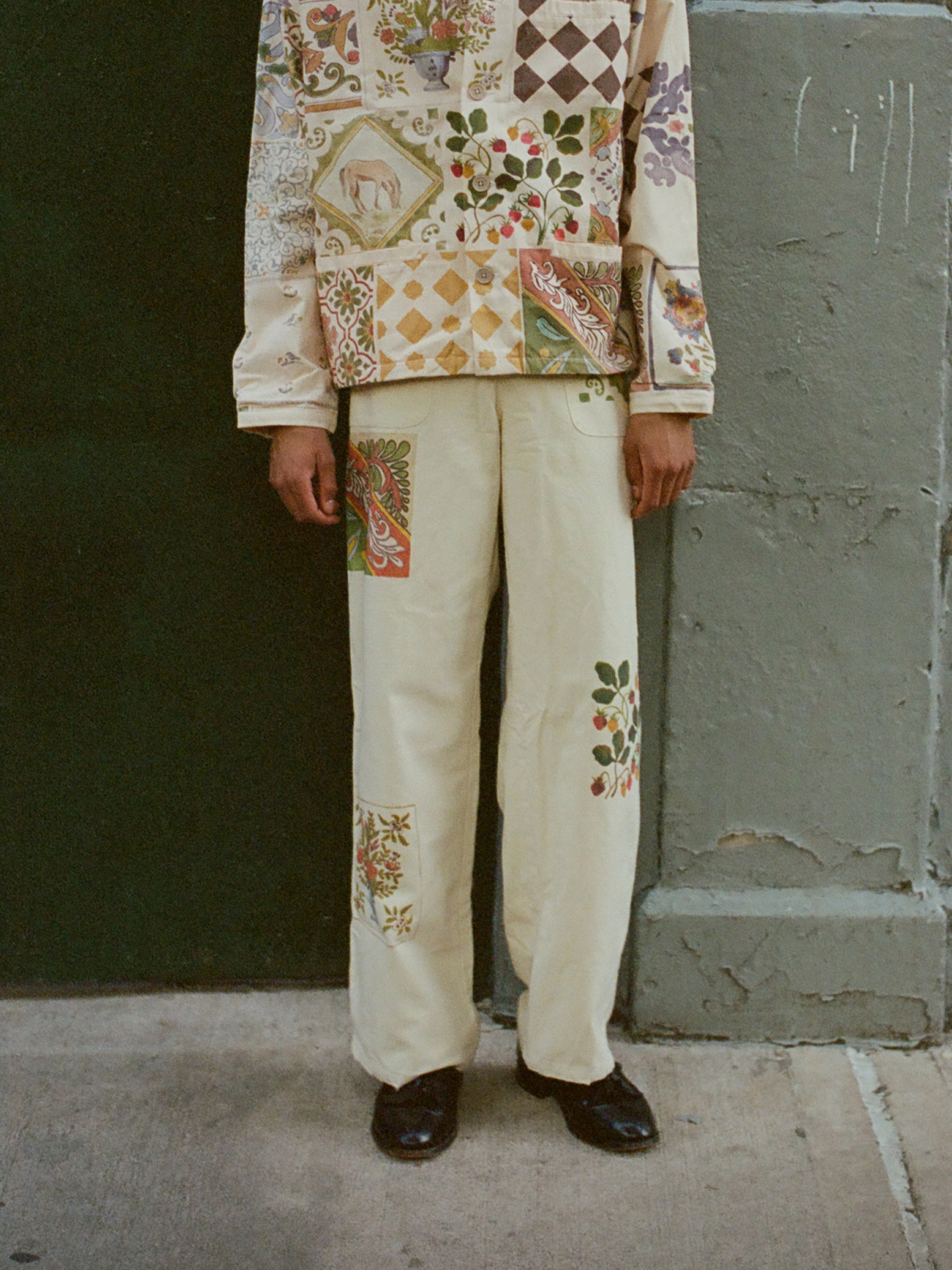 Against a green and gray wall, a person in vibrant Indo-Aryan designs, including FOUND's Mosaic Pants, showcases a matching patterned outfit.