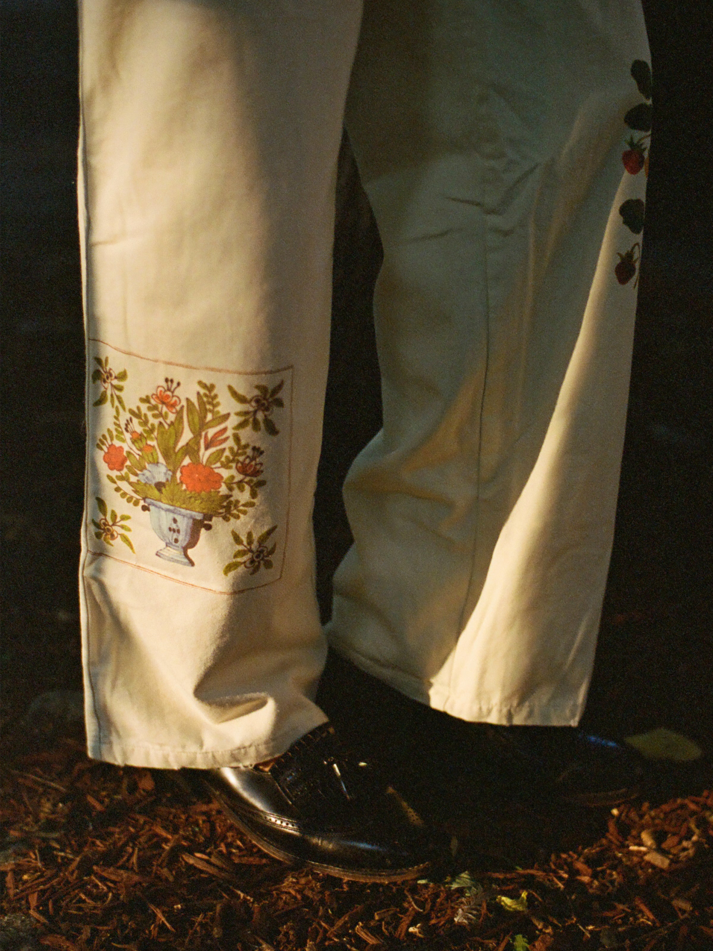A person stands on a leaf-covered ground wearing FOUND’s Mosaic Pants with floral embroidery and black shoes, combining unisex appeal with subtle Indo-Aryan patterns.