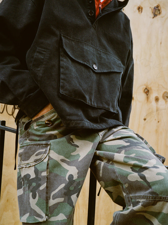 A person stands against a wooden background, wearing a black jacket with a large front pocket and FOUND Camouflage Twill Utility Cargo Pants.